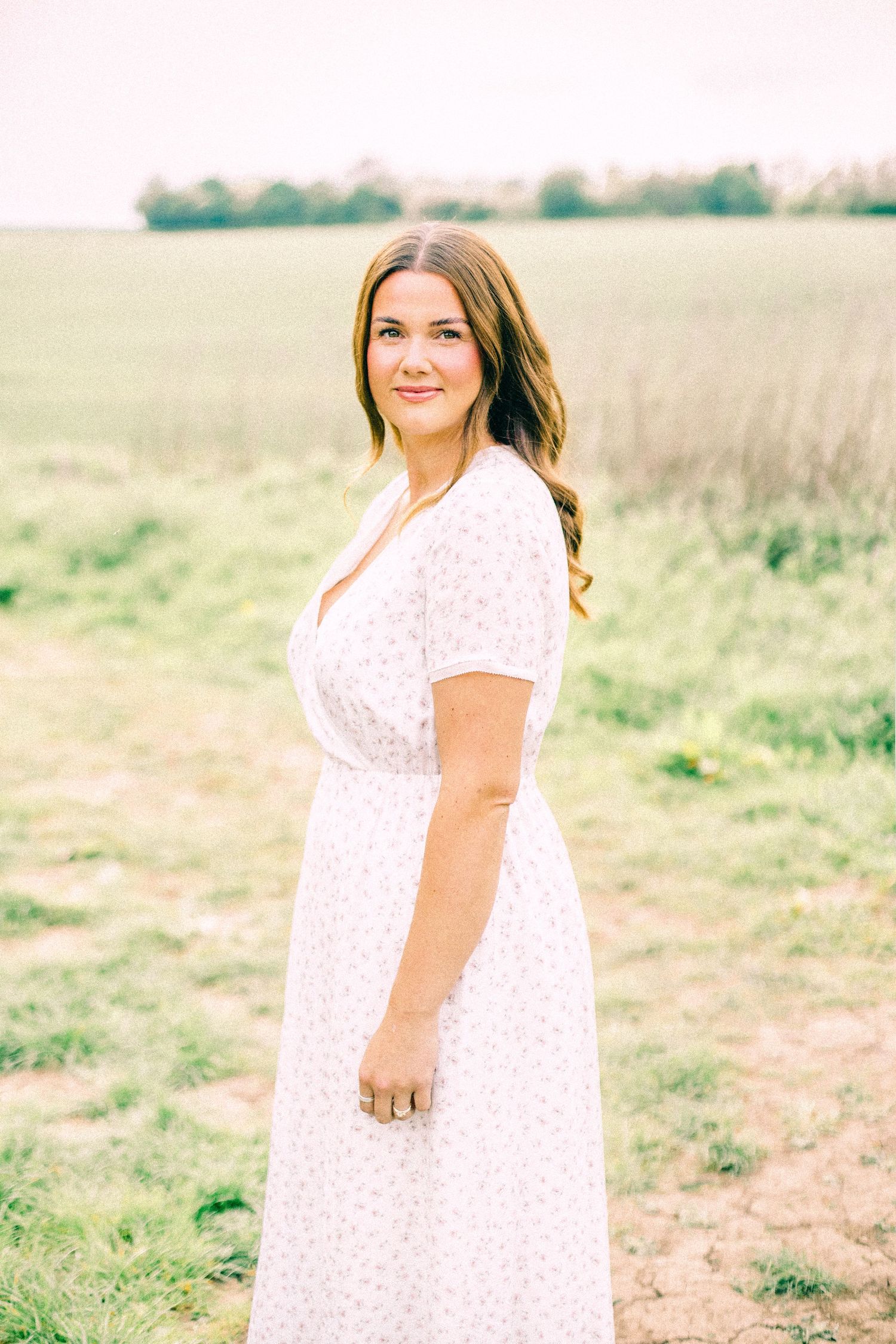 Woman with long auburn hair smiling in a white floral wrap dress, standing in a green grassy field at soft golden hour.