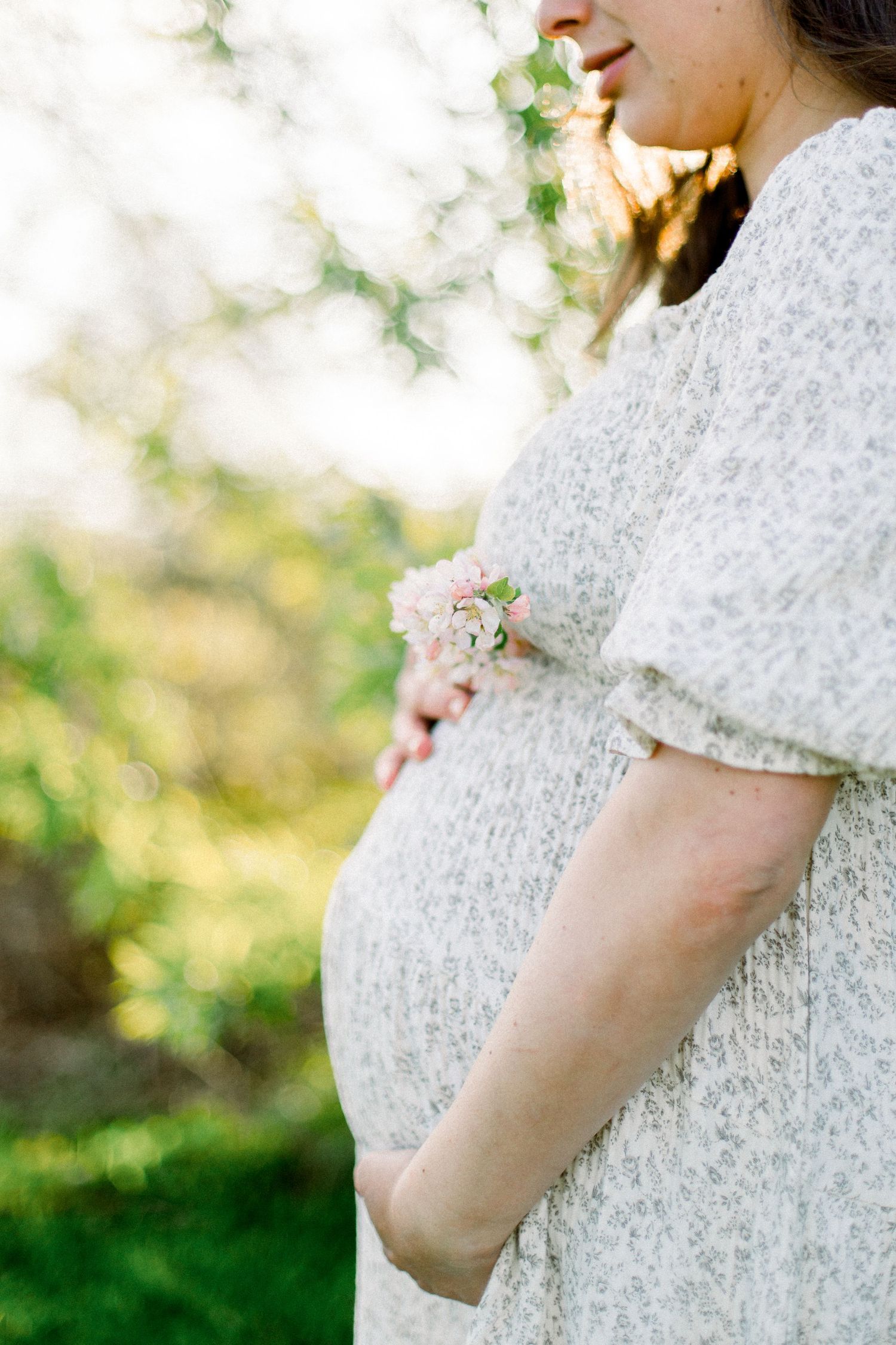 Side profile of a pregnant woman in a white floral dress cradling her baby bump while holding small pink flowers.