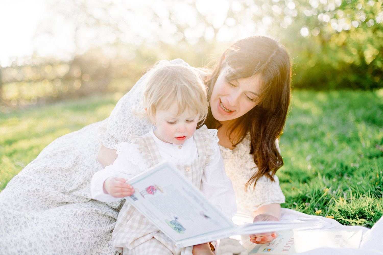 A mother and toddler sit on grass reading a picture book together in warm golden sunlight outdoors.