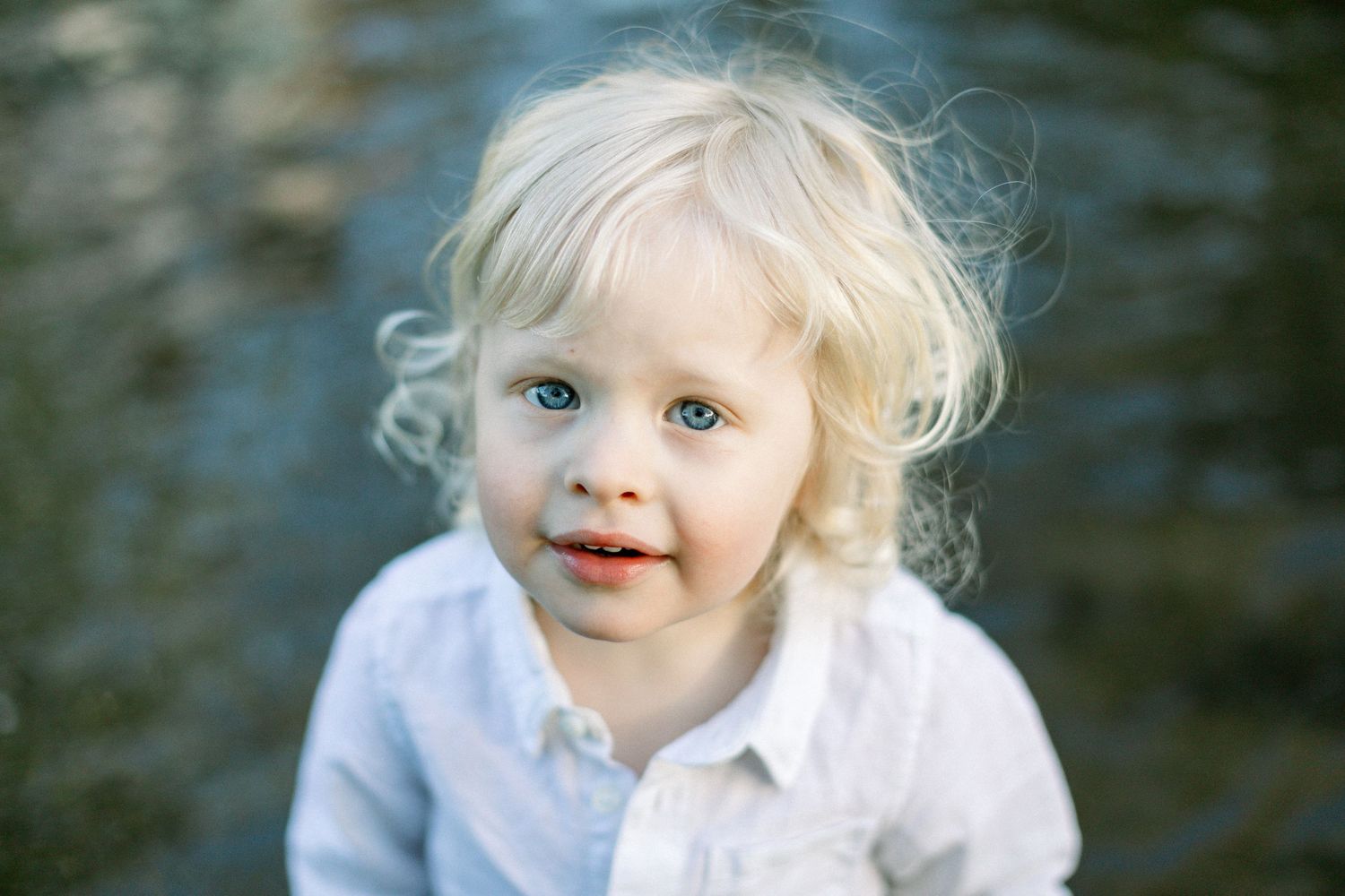 Adorable blonde toddler with blue eyes and curly hair wearing a white shirt, smiling near water outdoors.