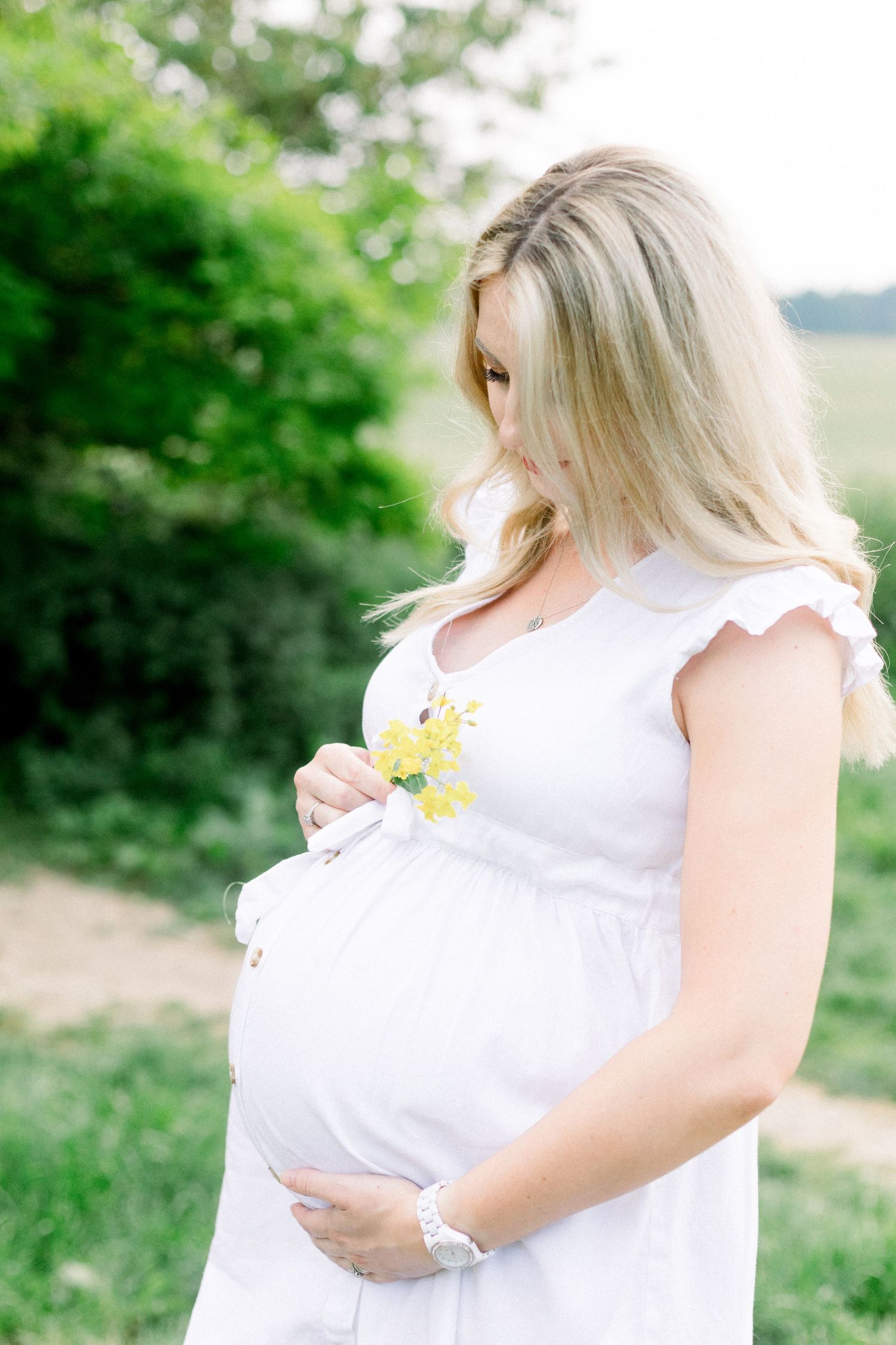 Pregnant woman in white dress holding baby bump outdoors, with yellow flowers and lush green trees in background.