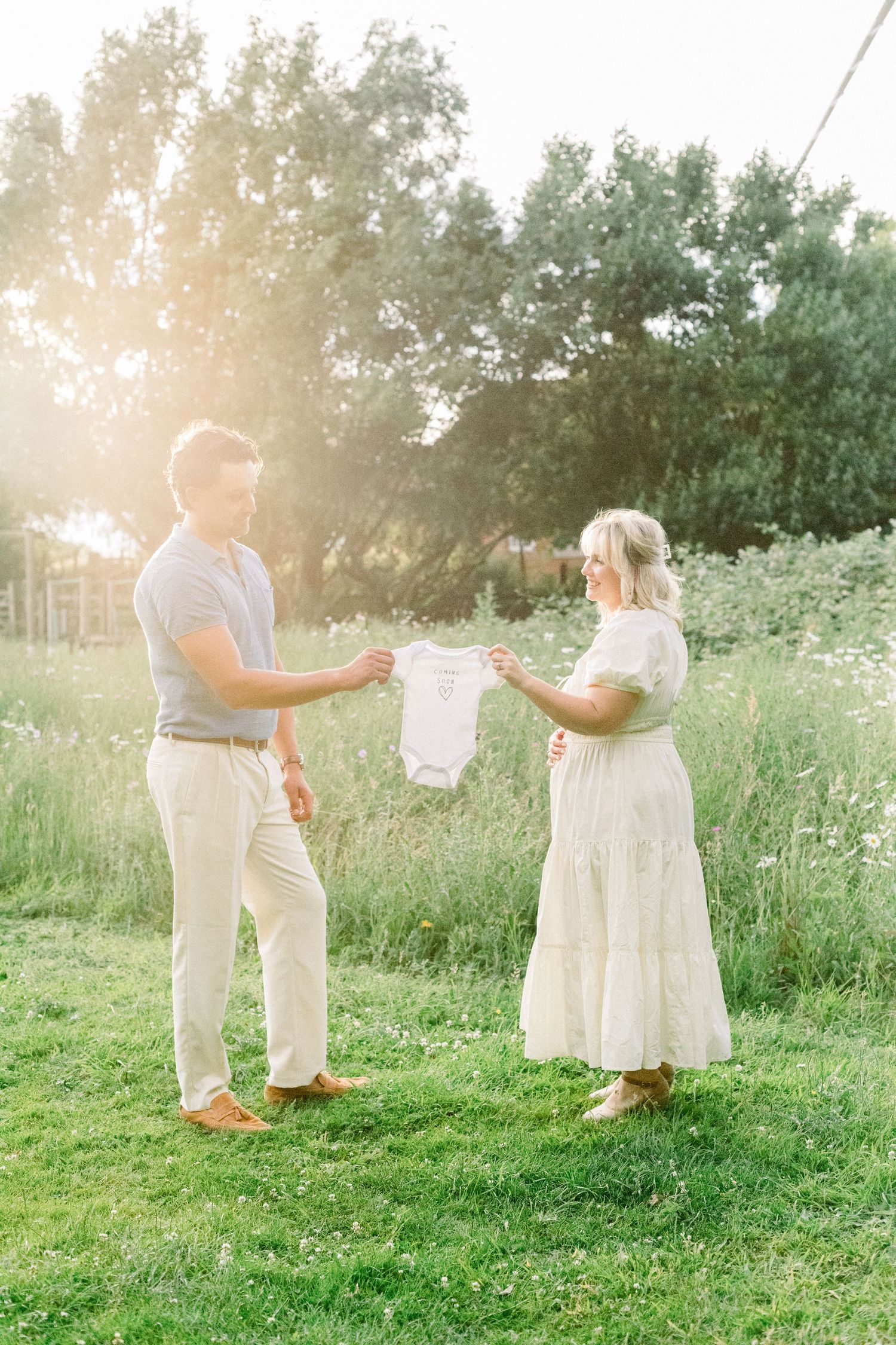 Expectant couple holding a baby onesie in a sunlit meadow, woman in white dress cradling her bump during maternity photoshoot.