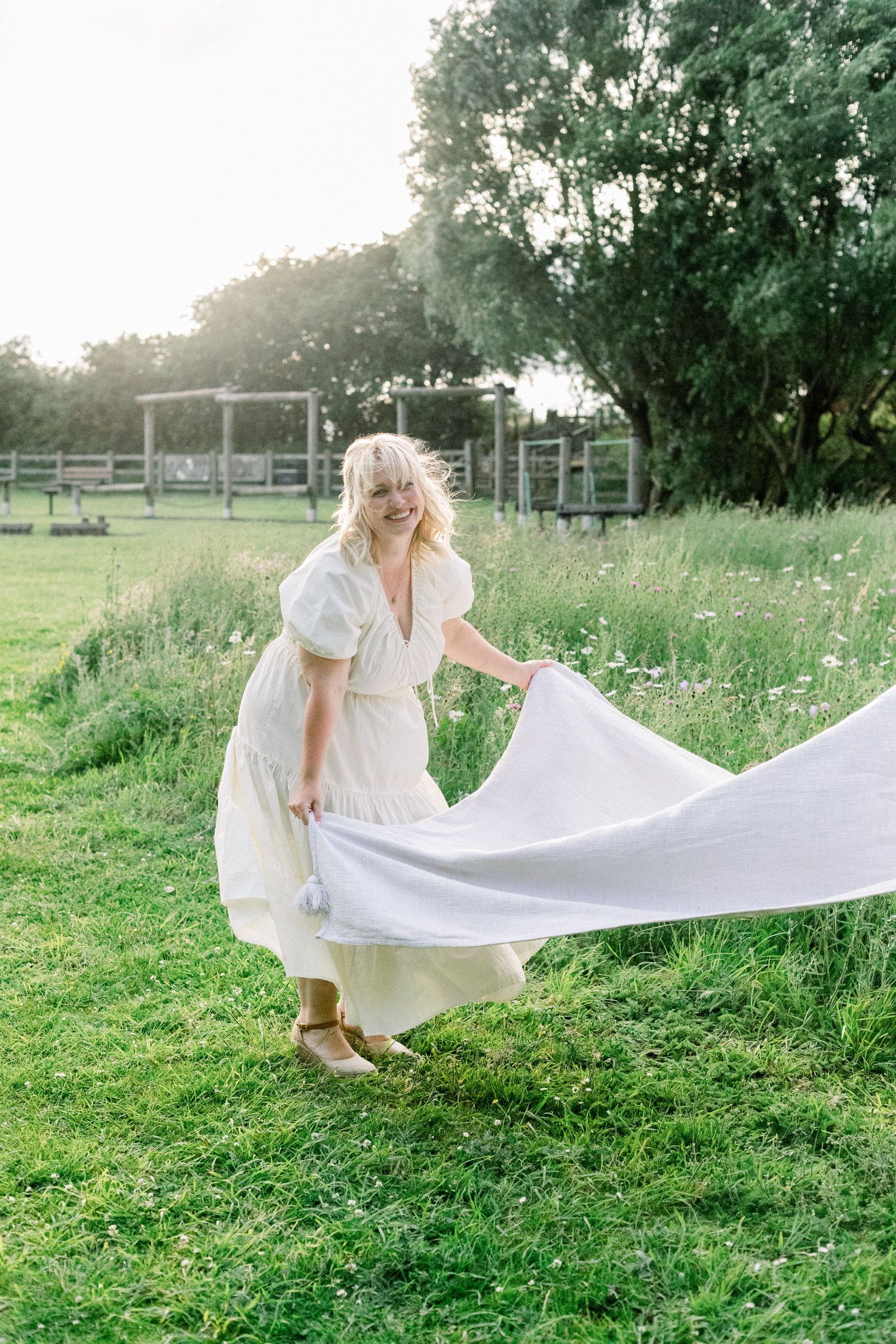 A smiling woman with blonde hair in a white dress holds a large white blanket while walking through a sunny green meadow.