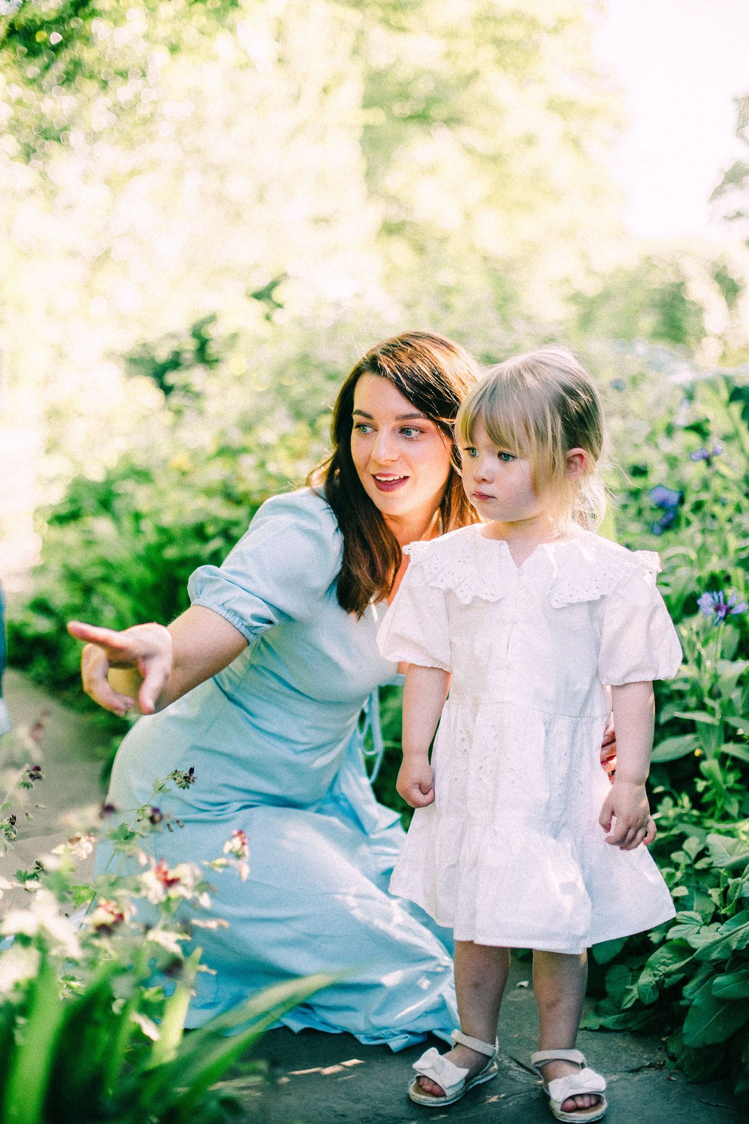 Woman in a light blue dress kneels beside a toddler in a white dress, pointing out colorful flowers in a sunlit garden.