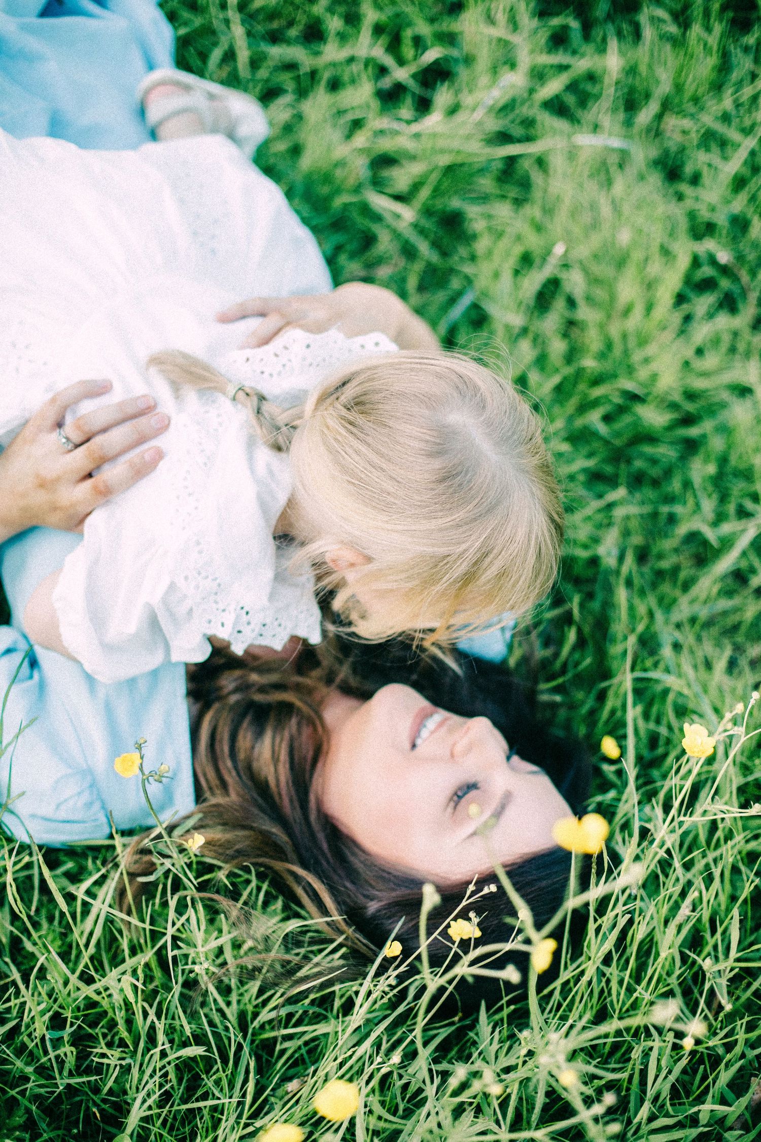 A smiling mother lies in green grass dotted with yellow buttercups while her blonde daughter in a white dress hugs her.