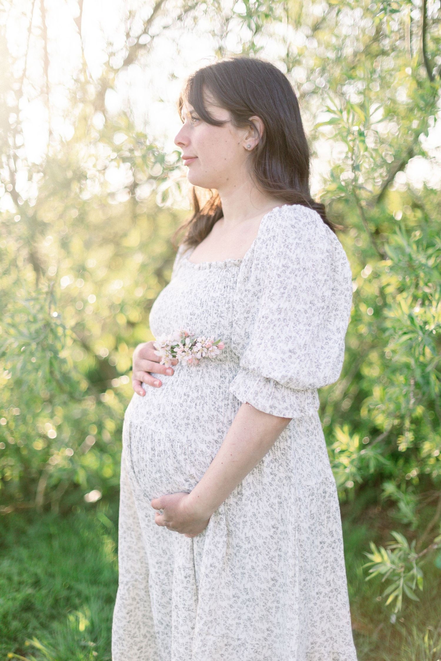 Pregnant woman in white floral dress holding baby bump outdoors in sunlit green garden during maternity photoshoot.