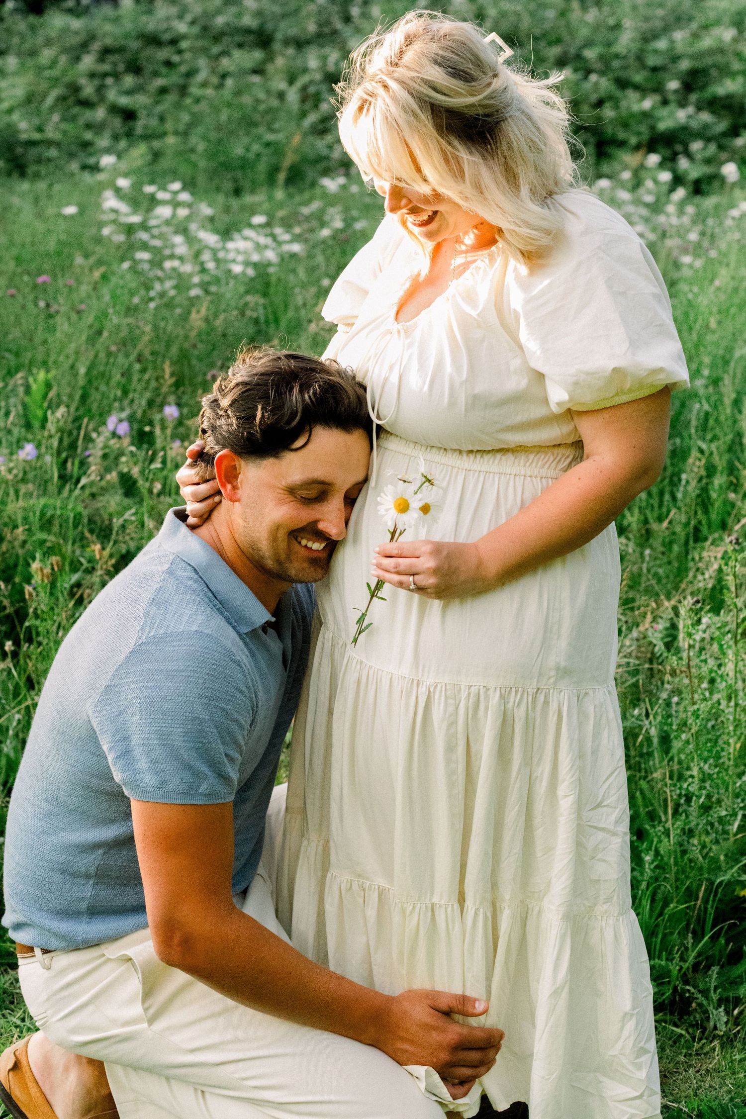 A smiling man kneels beside a pregnant woman in a white dress holding daisies in a lush green meadow.
