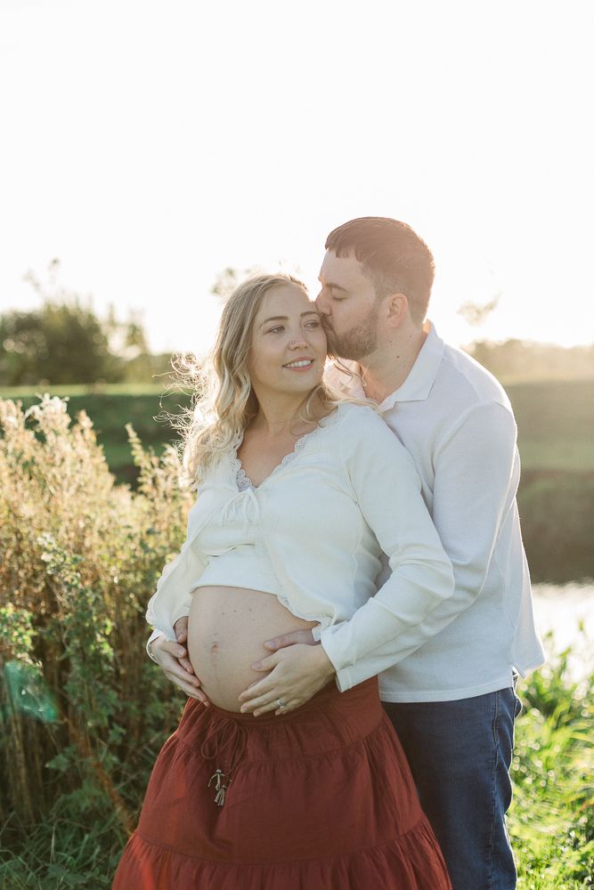 A couple shares a tender moment outdoors at golden hour, with the man kissing his pregnant partner's temple.