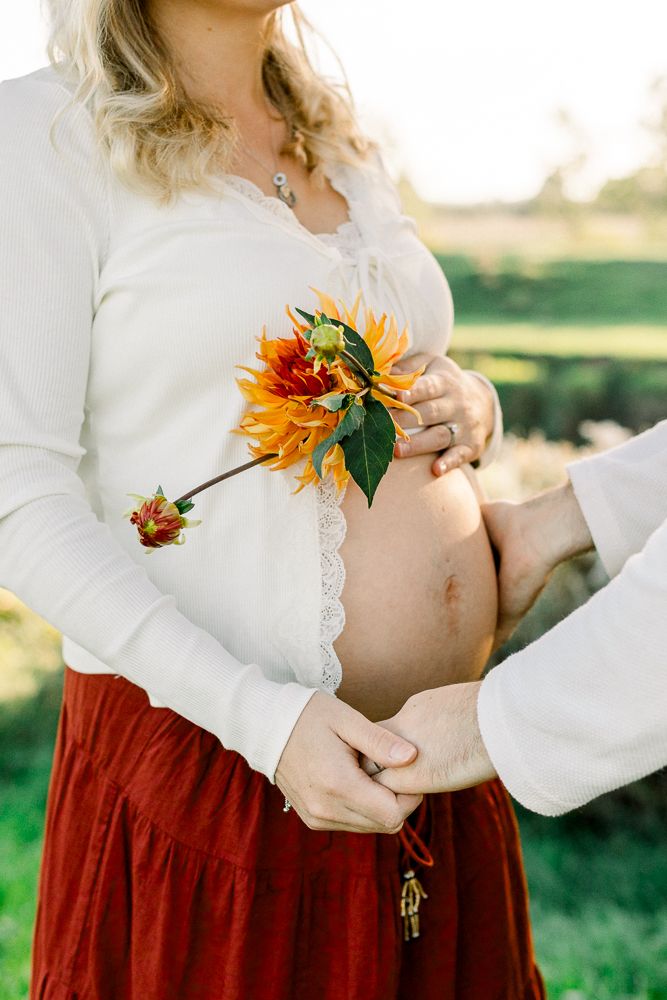 Pregnant woman in white top and red skirt holds a sunflower over her baby bump outdoors.
