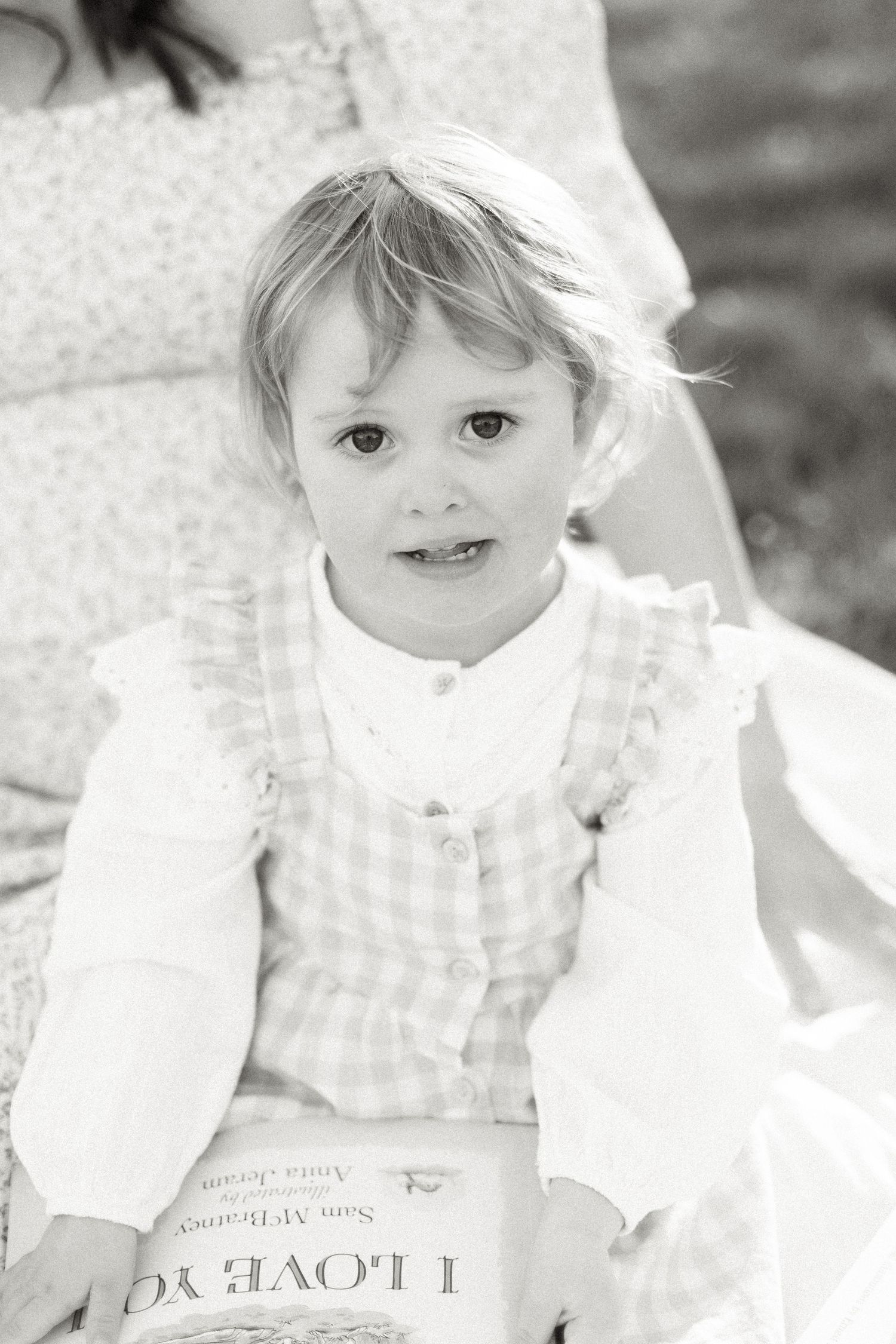 Black and white portrait of a blonde toddler girl in a gingham dress holding an I Love You book outdoors.