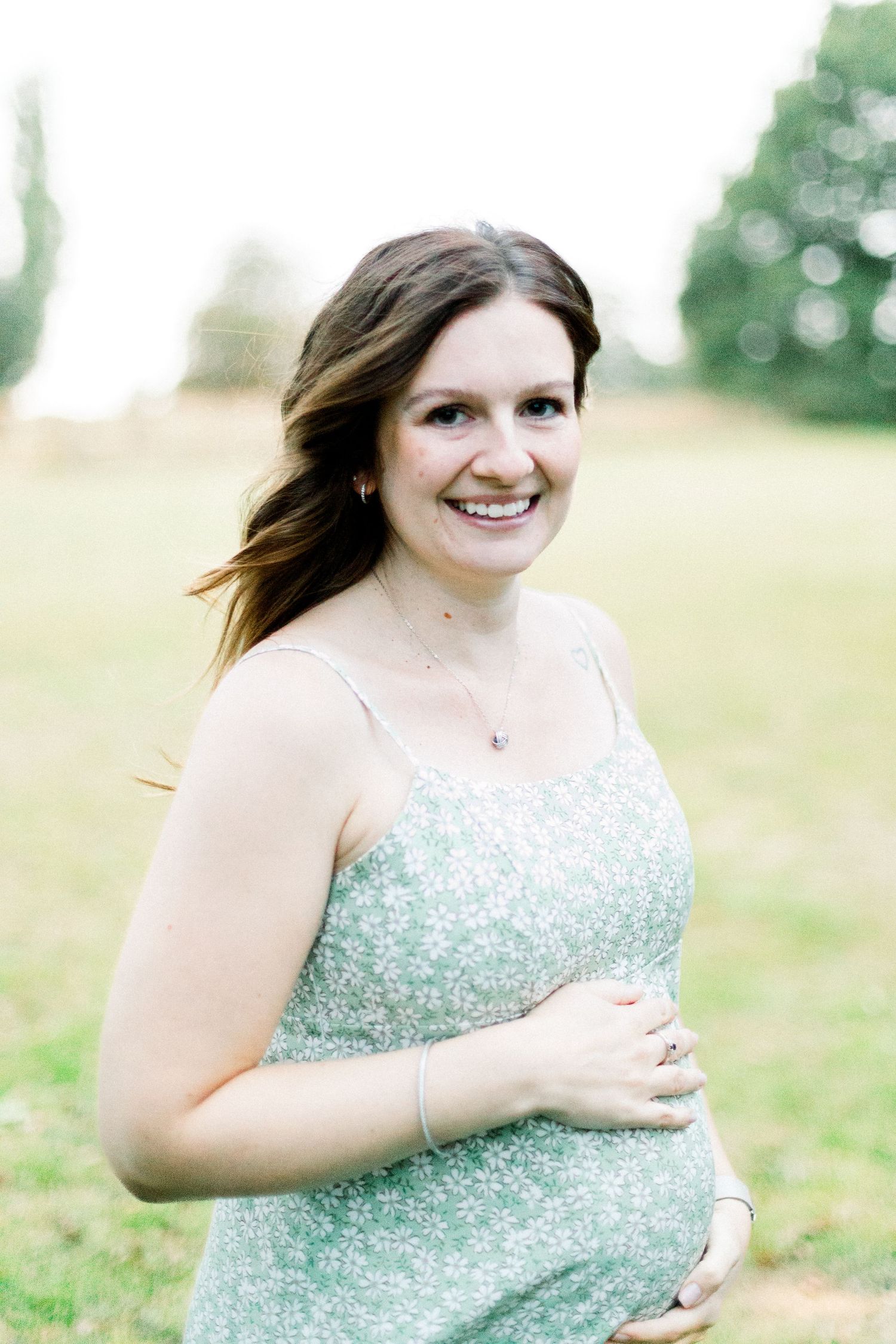 Smiling pregnant woman in a green floral sundress cradling her baby bump outdoors in a park setting.