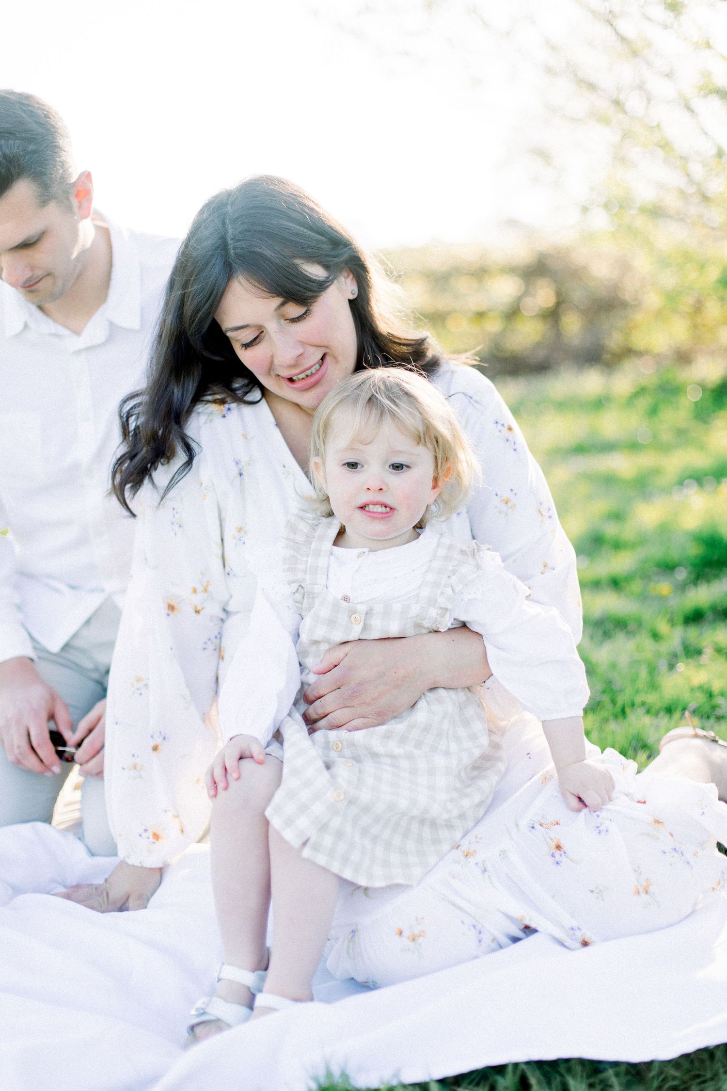 A mother in white hugs her toddler girl on a blanket outdoors during a bright spring family photo session.