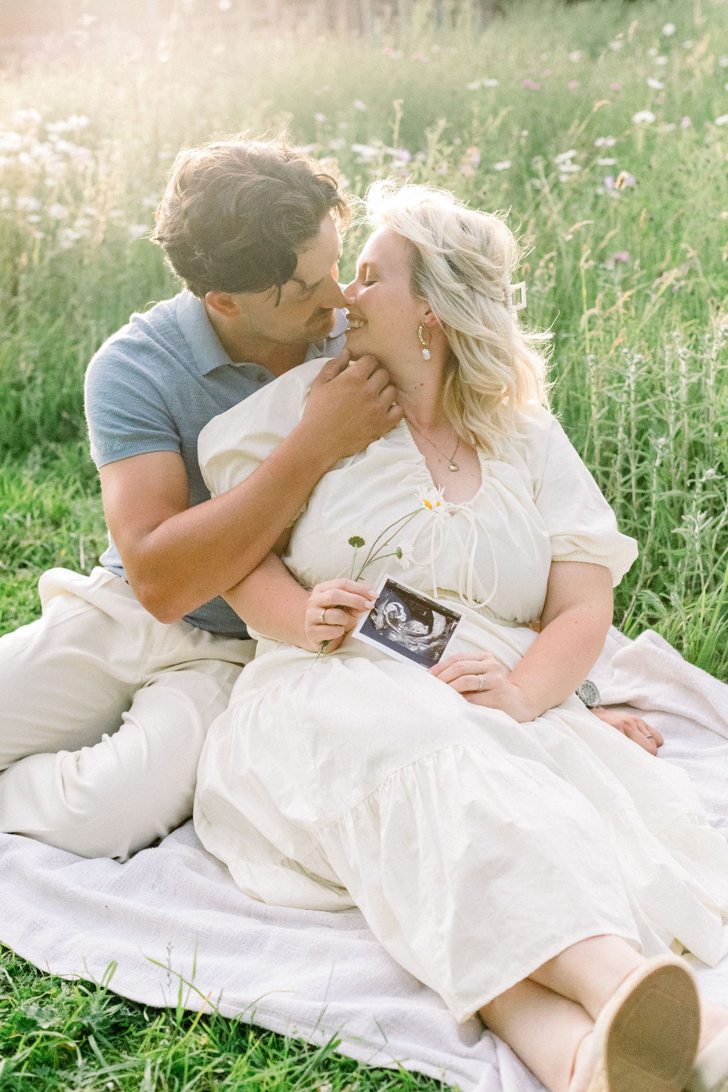 Couple embracing and kissing on a white blanket in a sunlit wildflower meadow while holding an ultrasound photo in a maternity announcement.