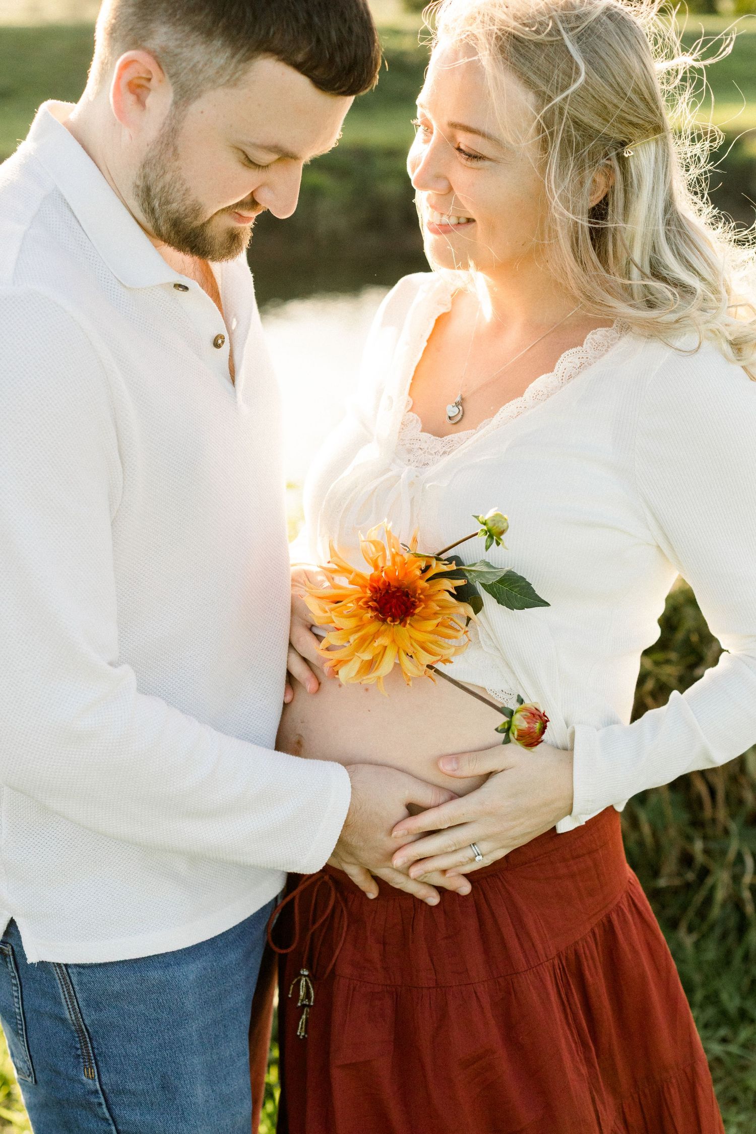 Couple tenderly cradling pregnant belly outdoors, holding yellow dahlia flower in warm golden sunlight.