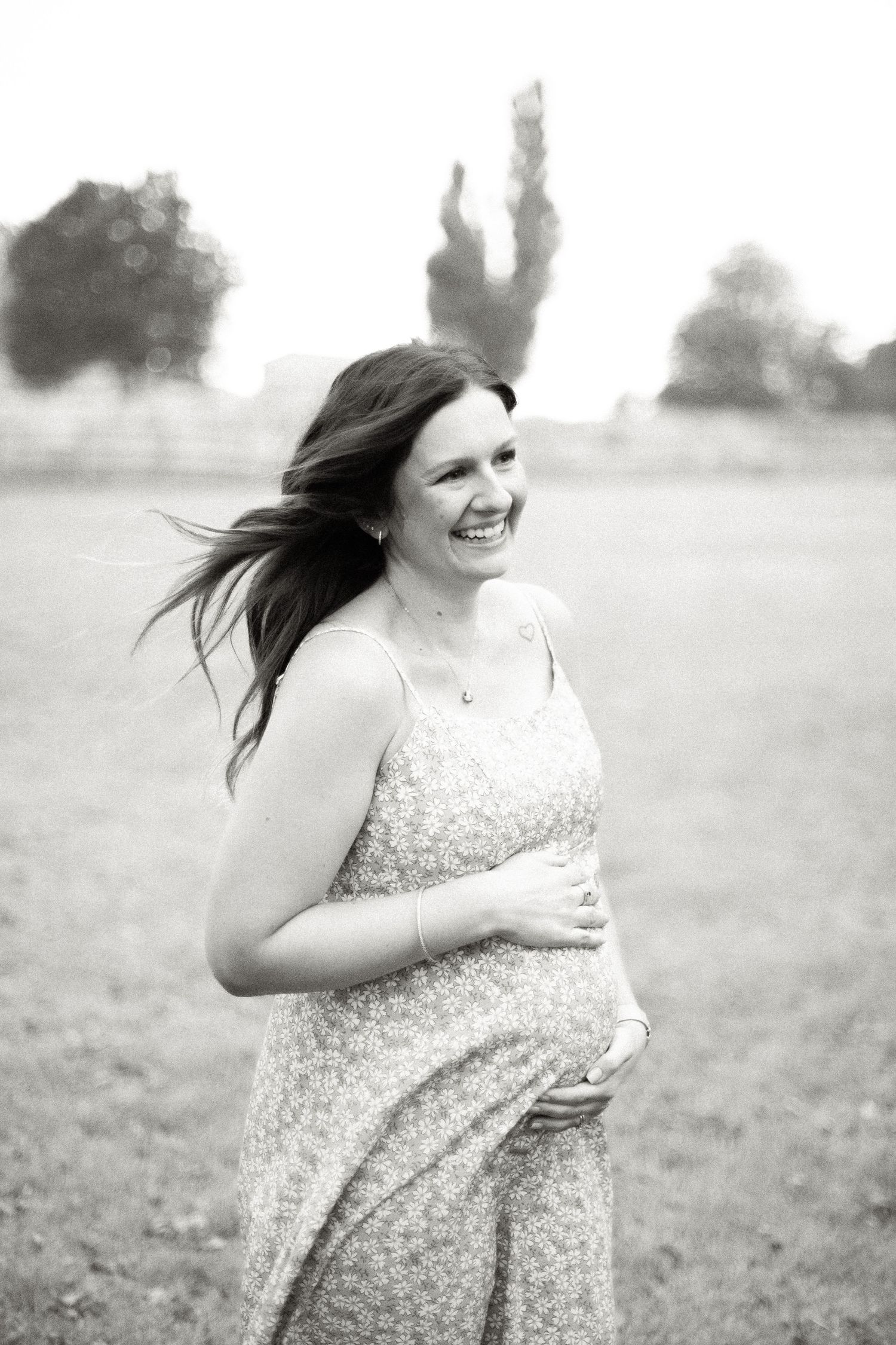 Smiling pregnant woman in floral dress holding baby bump outdoors in black and white maternity photo.