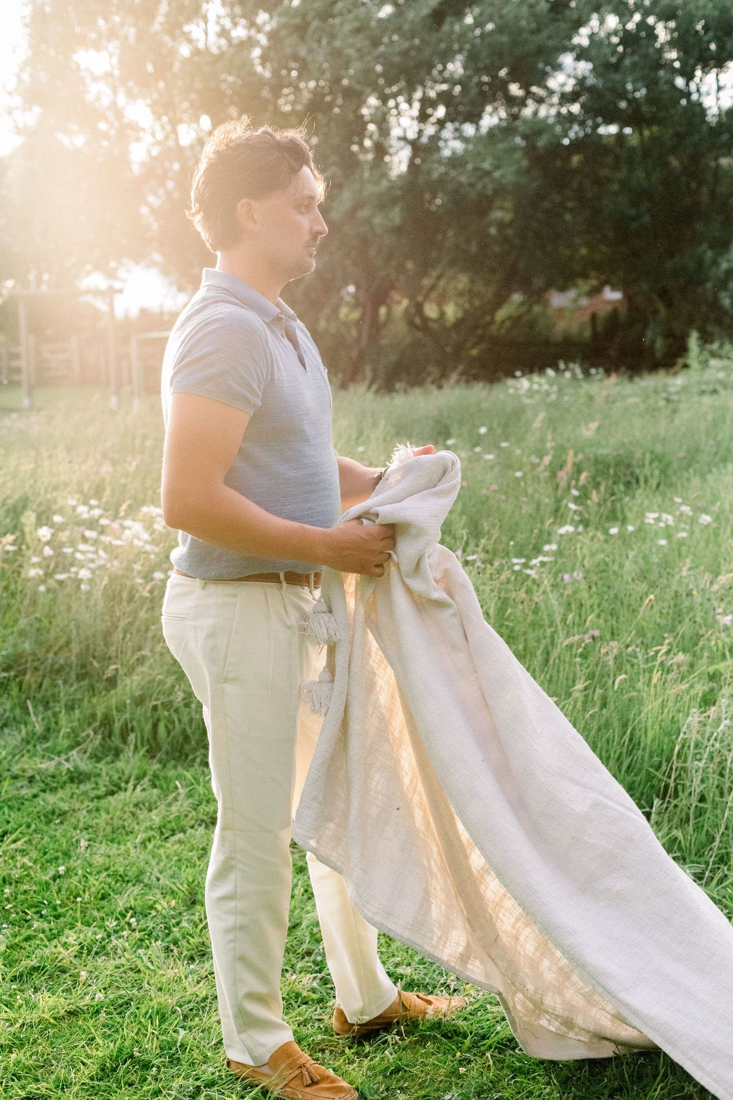 Man in a gray polo, khaki pants and tan shoes holds a large linen blanket in a sunlit wildflower meadow.