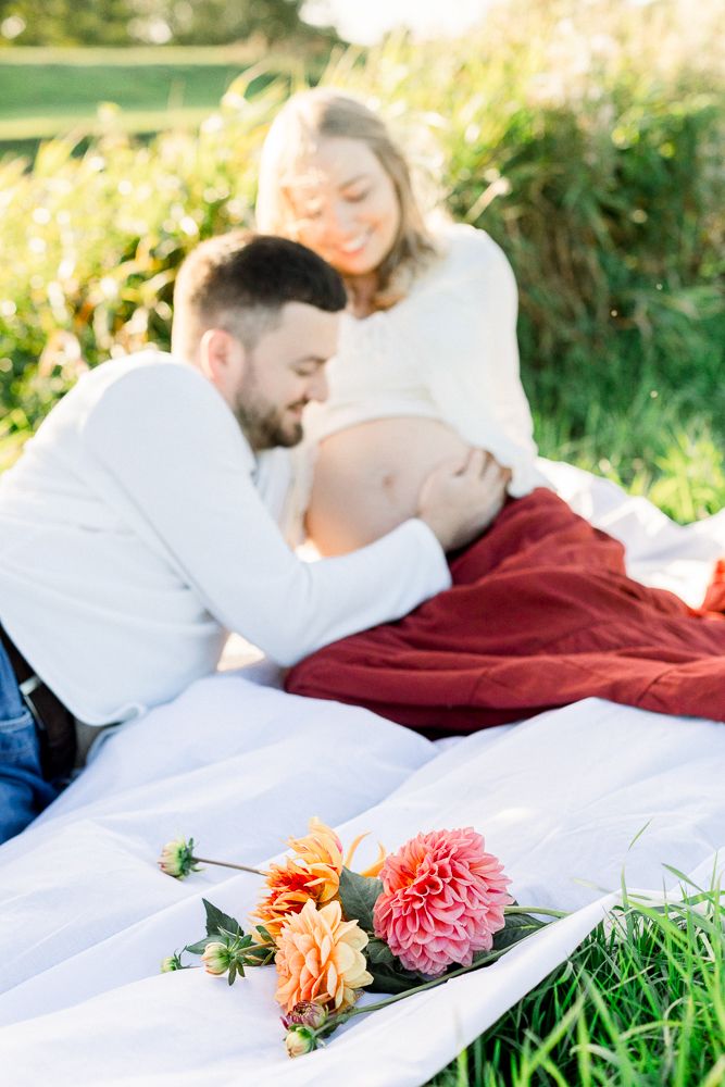 Couple sharing a tender maternity moment outdoors on a white blanket with colorful dahlias in the foreground.