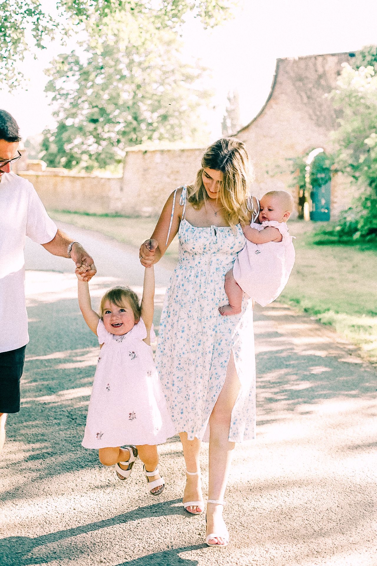 A smiling woman in a floral dress carries a baby while a toddler in a pink dress swings between her and a man on a sunny path.