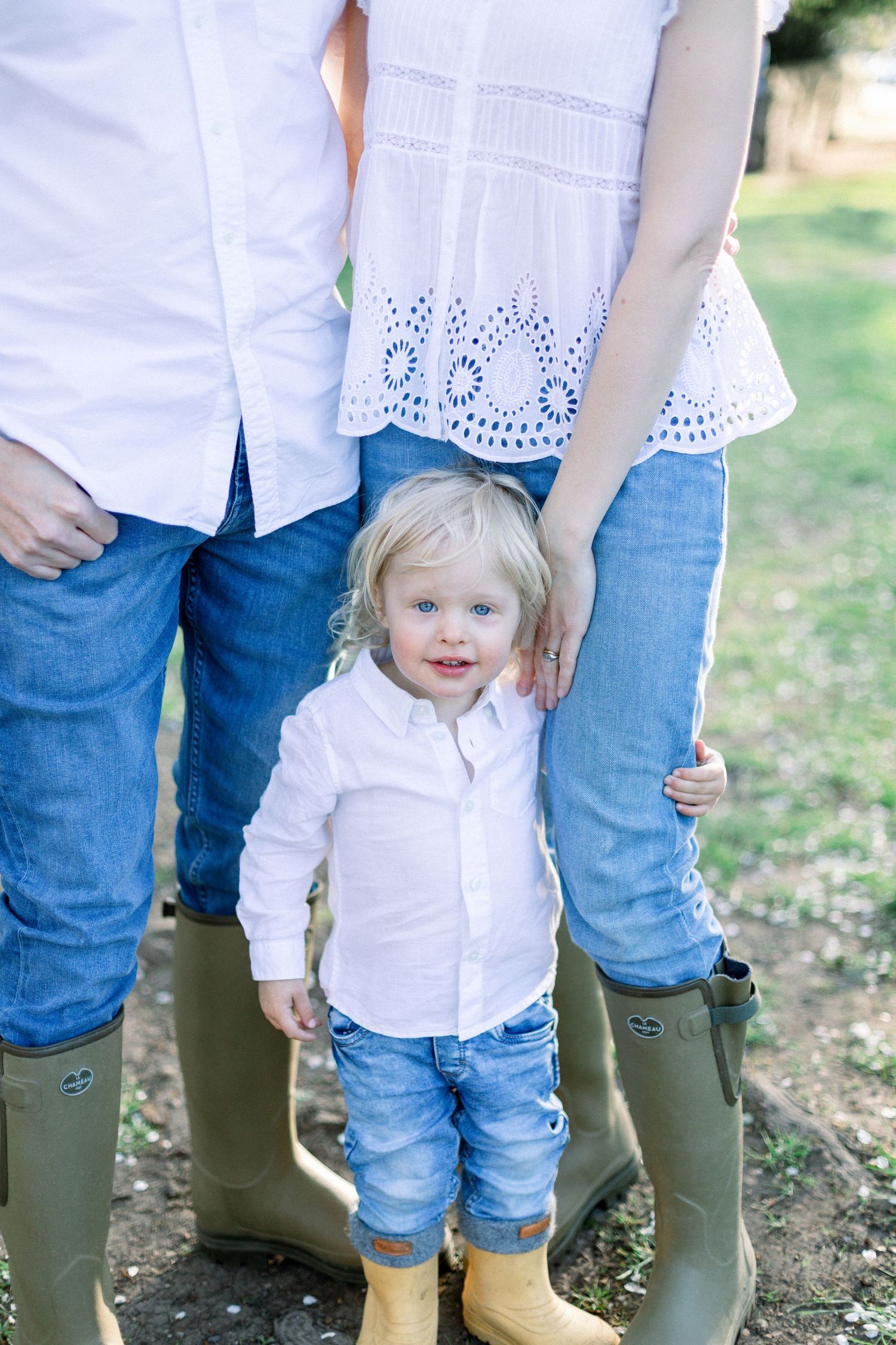 Toddler boy in white shirt and jeans stands between parents wearing denim and rain boots outdoors.