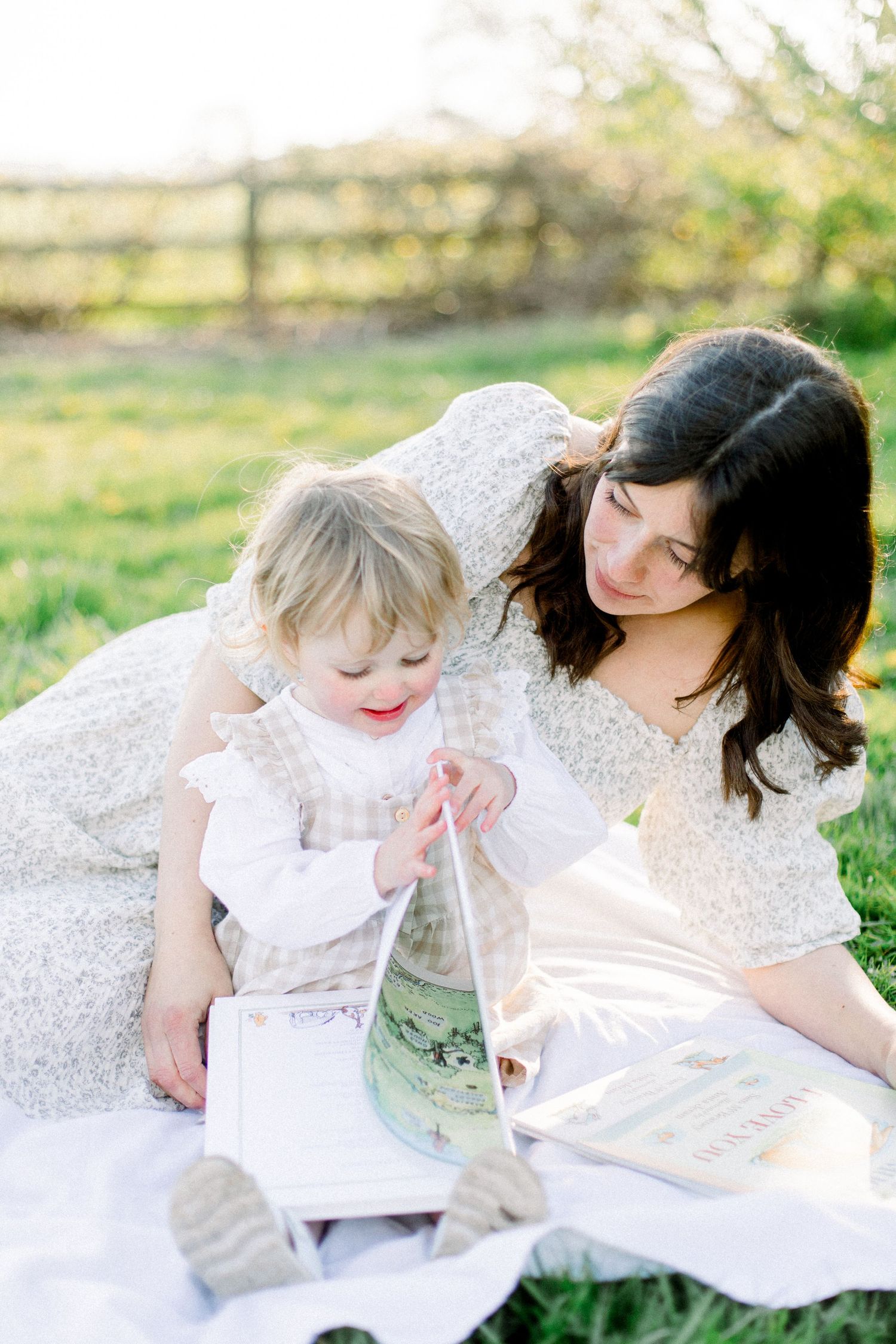 A dark-haired mother and blonde toddler read a book together on a blanket in a sunny green field.