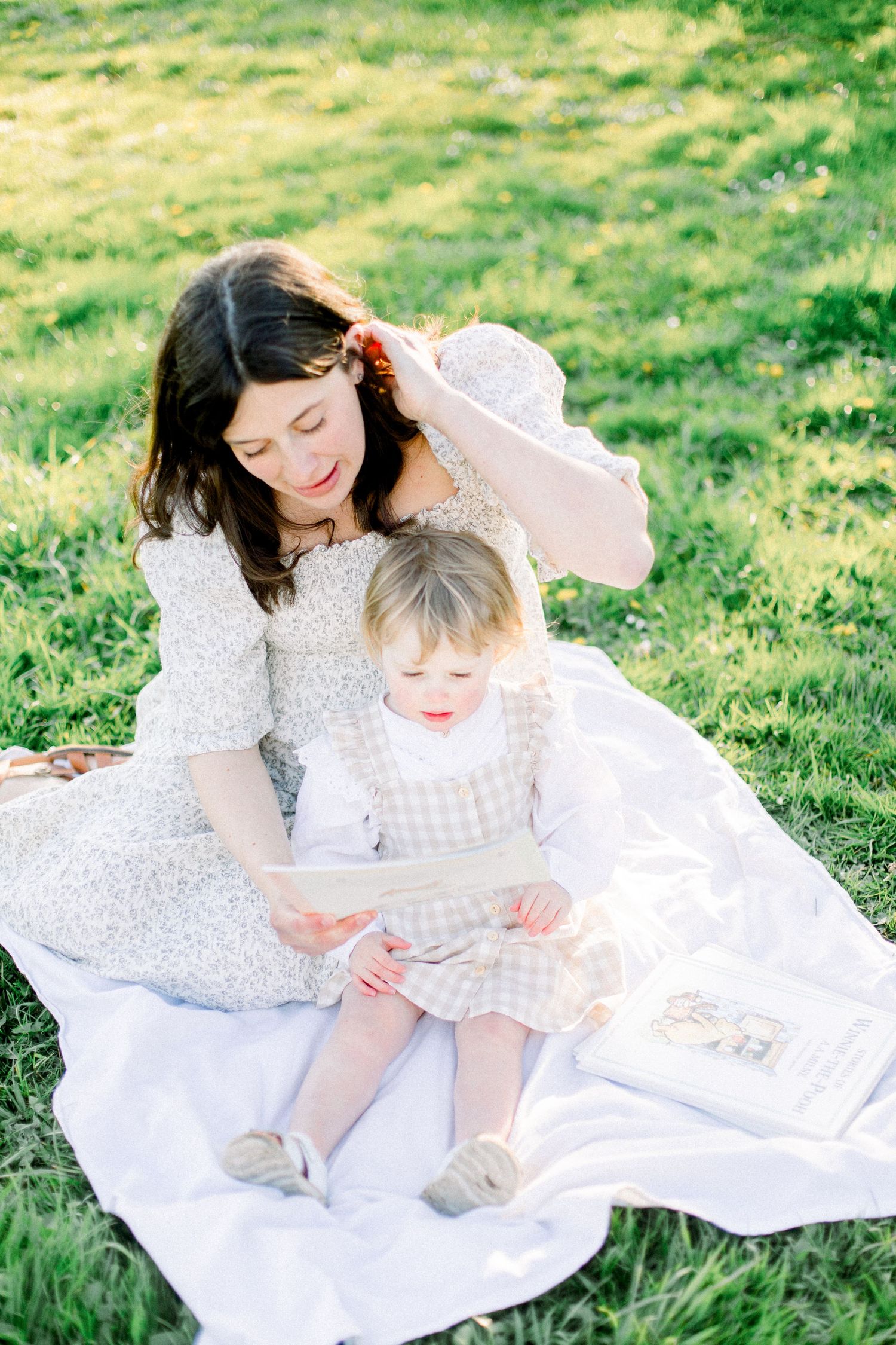 Mother and toddler daughter sit on white blanket in sunny park, reading a book together in matching floral outfits.