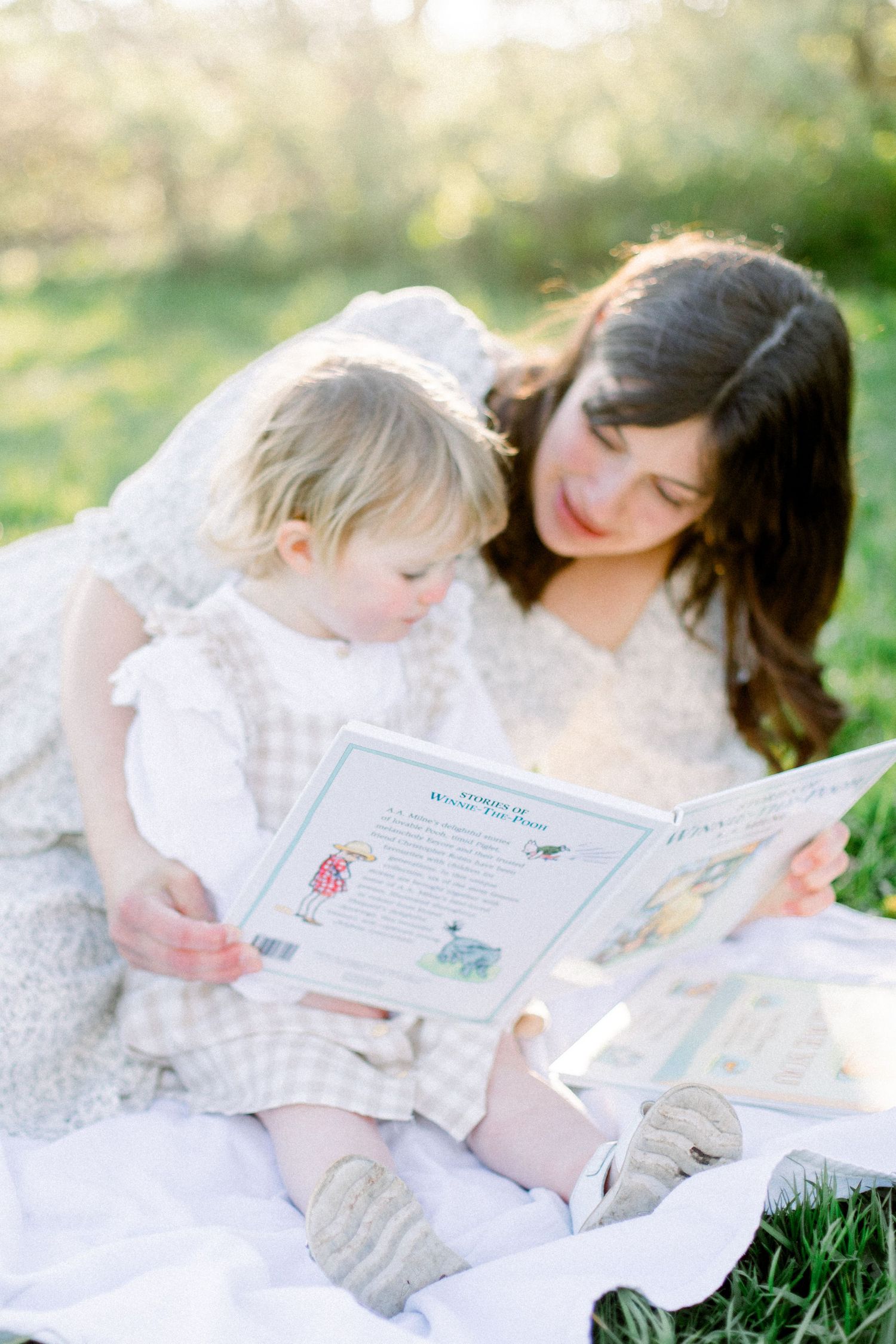 Mother and toddler reading Winnie the Pooh book together outdoors on a blanket in a sunlit garden.