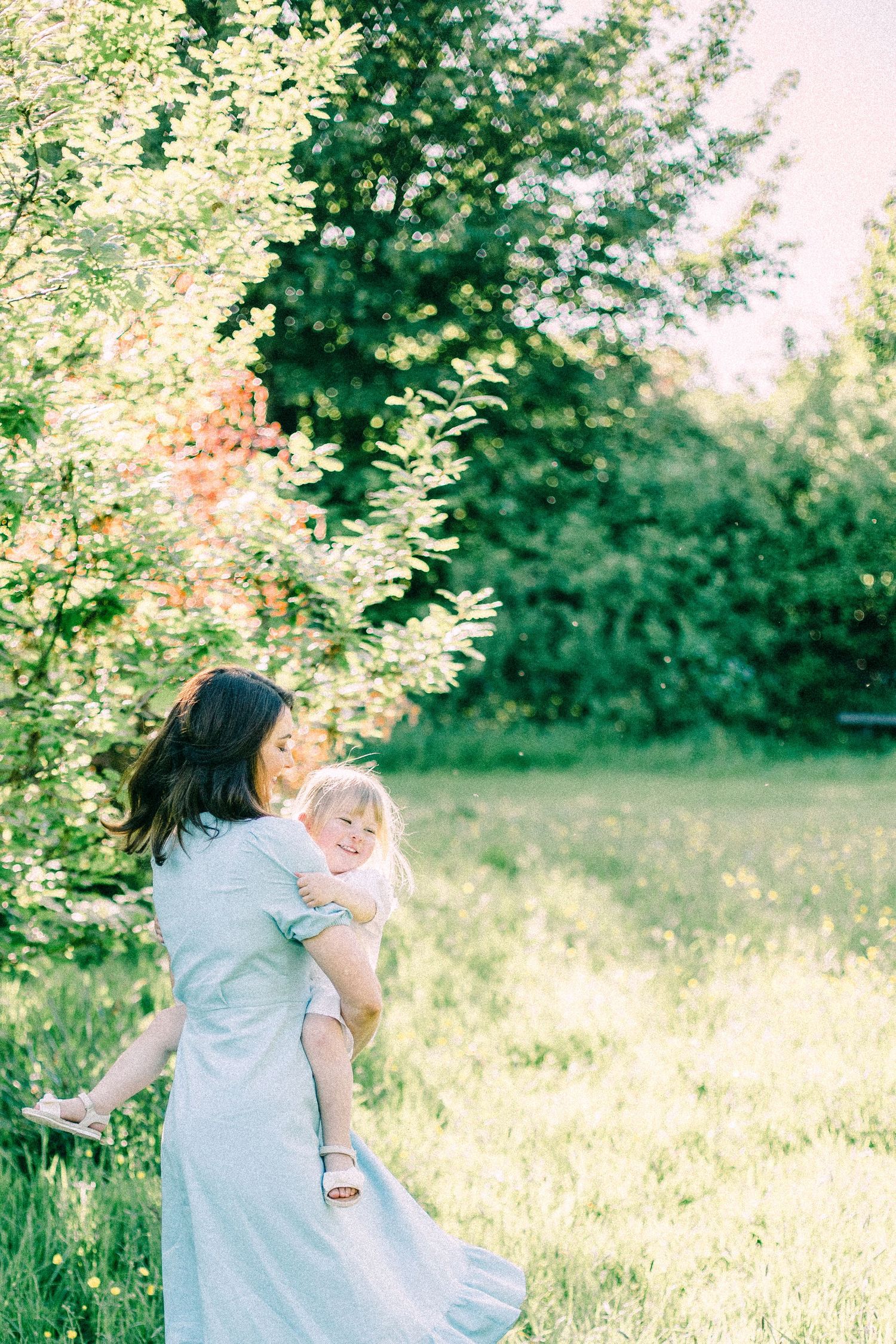 A woman in a light blue dress holds a smiling toddler in her arms while standing in a sunlit green meadow beside blooming trees.