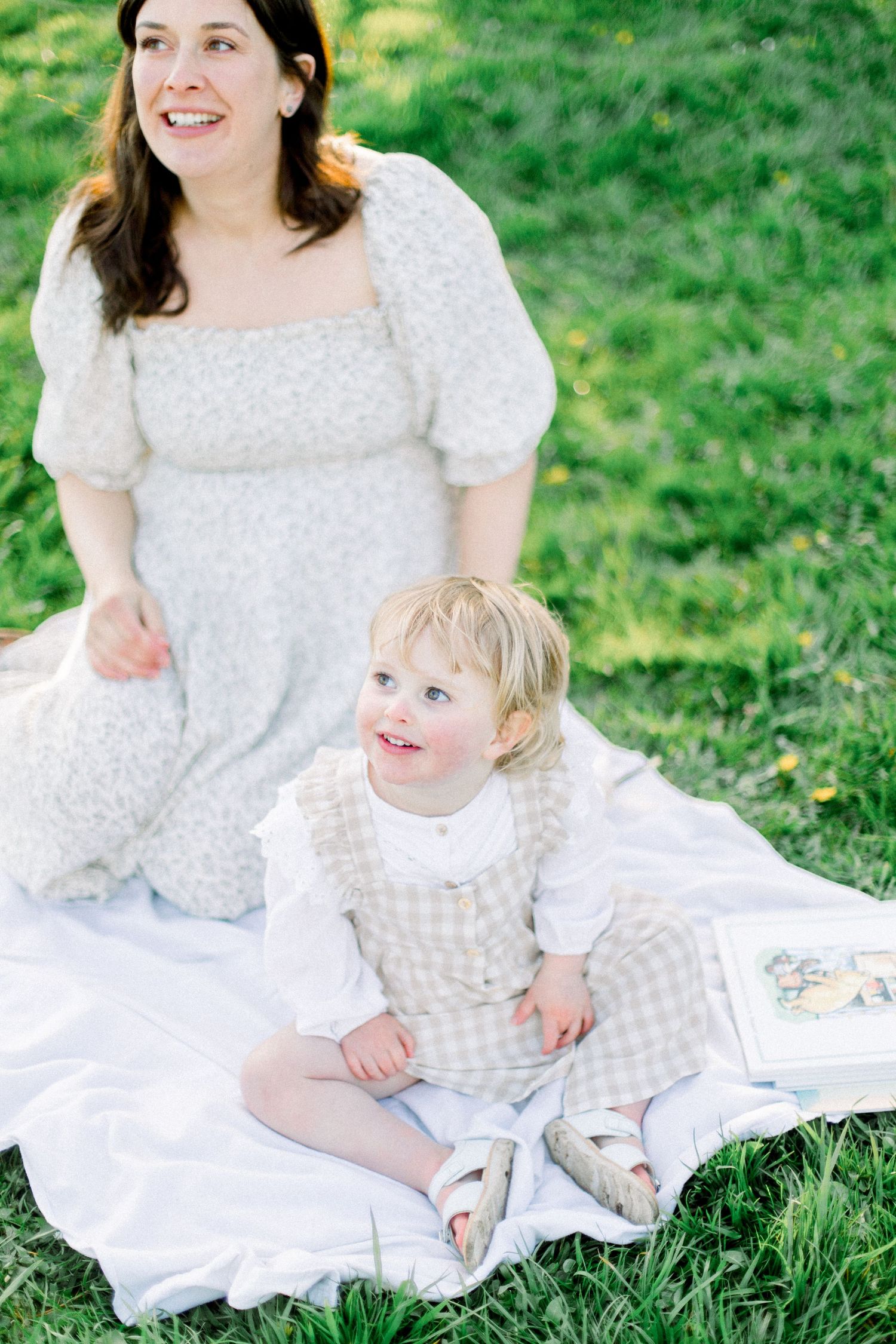 A woman in a white dress sits with a toddler on a blanket in green grass.