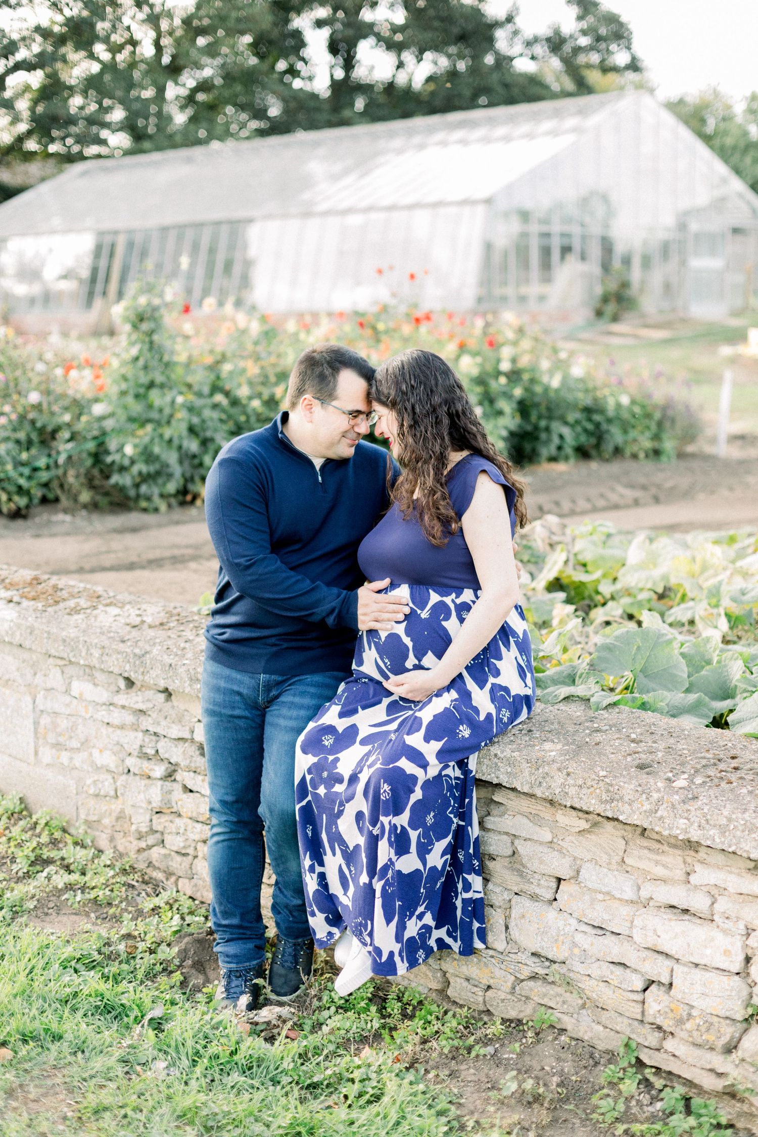 Expecting couple touching foreheads as the woman in a blue floral dress sits on a stone wall near a greenhouse and flower garden.