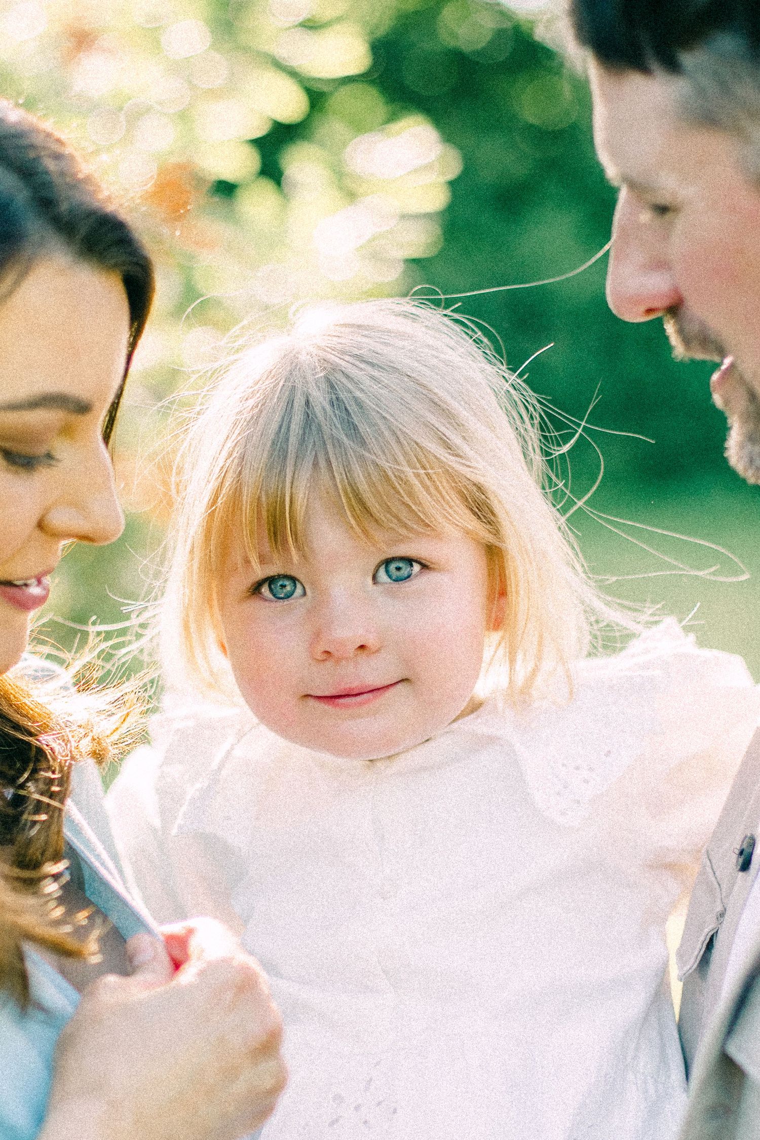 Smiling toddler girl with blonde hair and blue eyes in a white dress, held by her parents in warm golden sunlight outdoors.