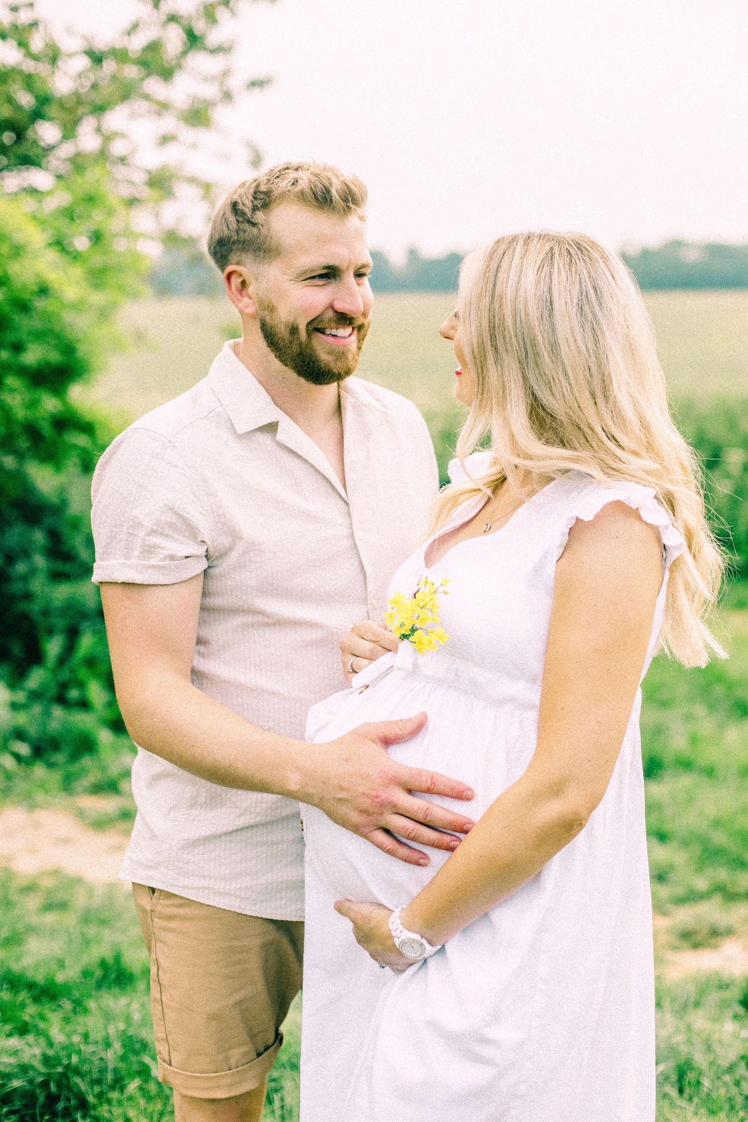 Smiling bearded man gently touches the baby bump of his pregnant partner in a white dress holding yellow flowers in a sunny green field.