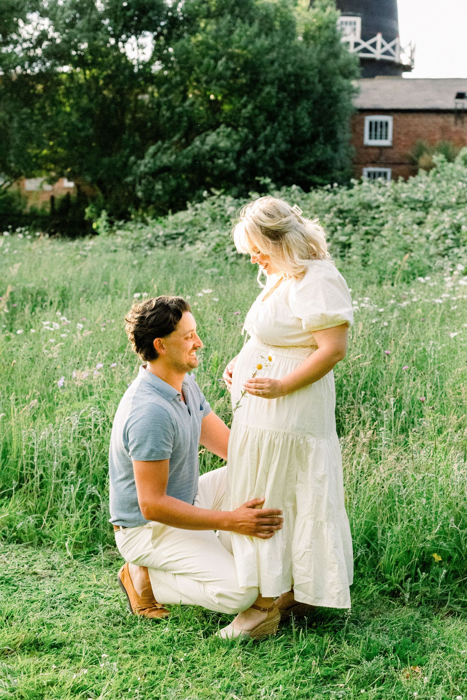 A man kneels in a meadow, smiling up at a pregnant woman in a white dress during a maternity photoshoot.