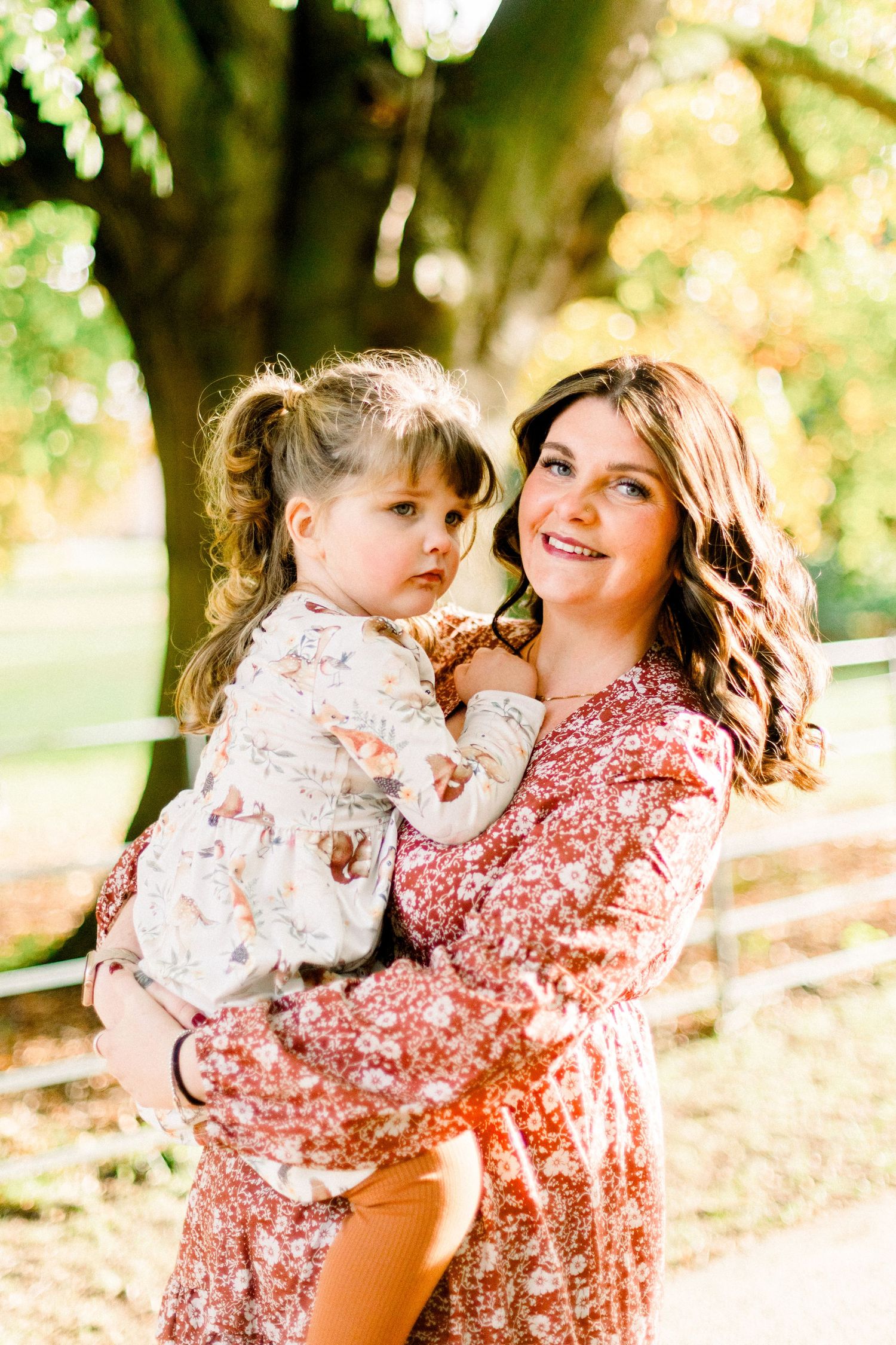A smiling woman in a floral dress holds a young toddler girl outdoors in a sunlit park setting.