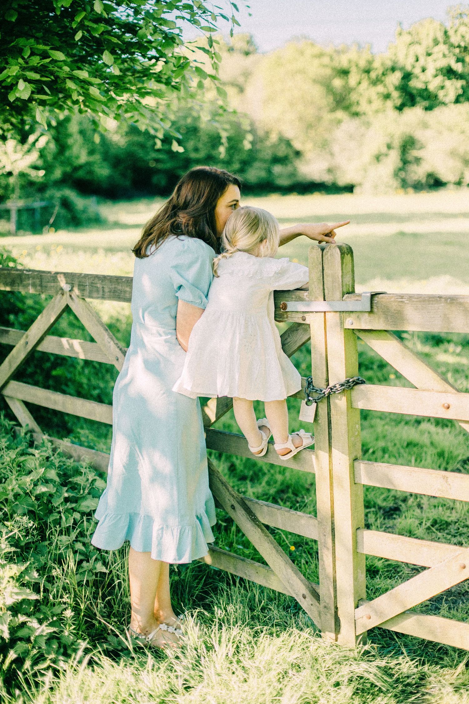 A woman in a light blue dress lifts a toddler in a white dress to see over a wooden farm gate while pointing across a sunny green meadow.