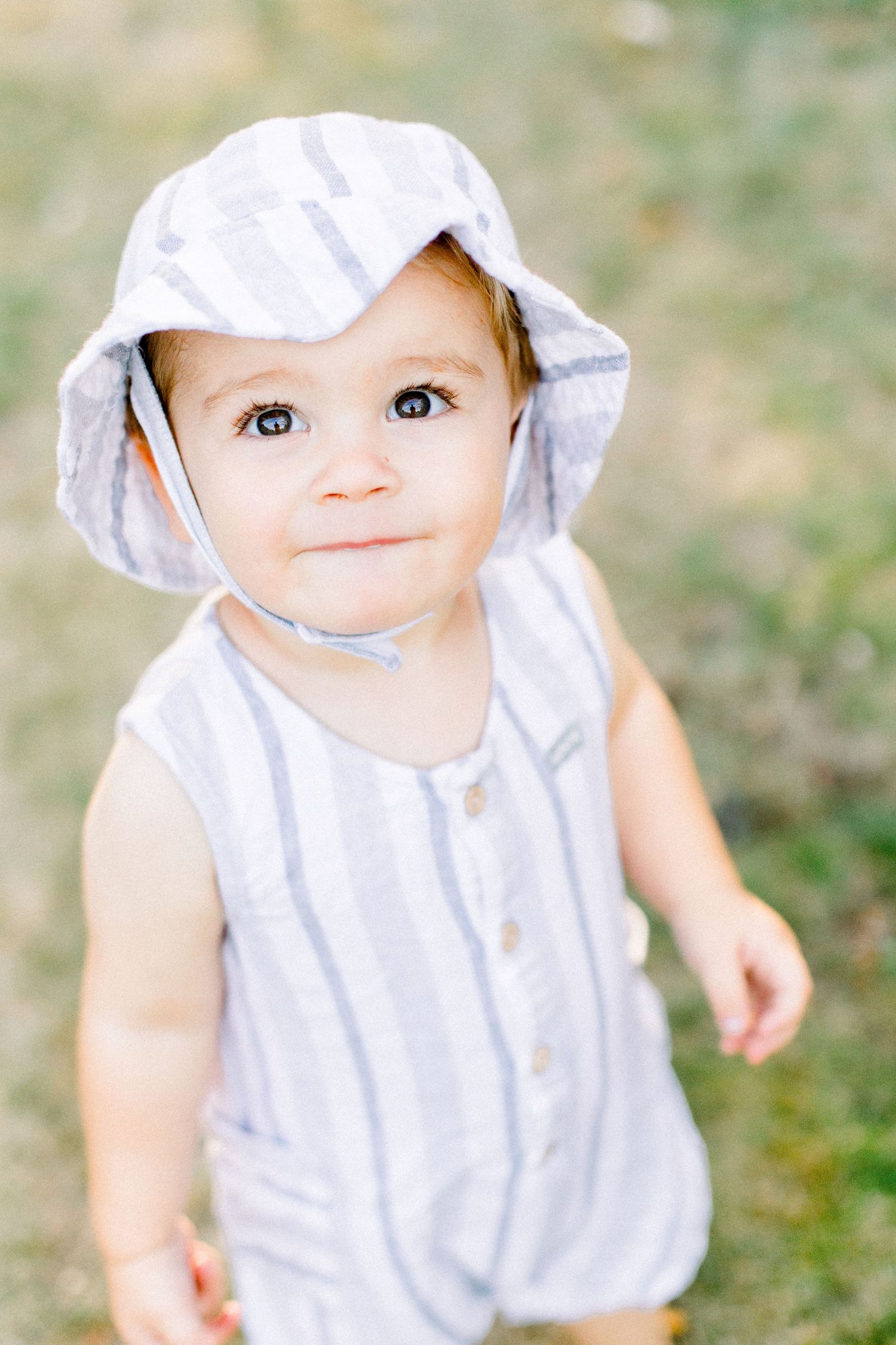 Adorable baby in striped romper and matching sun hat smiling outdoors on green grass in summer.