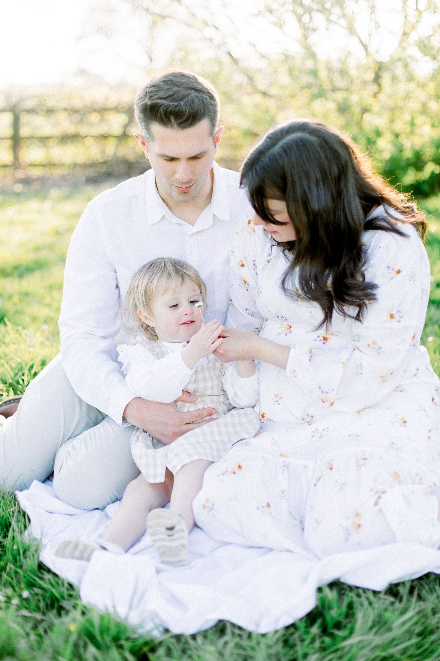A young family sits on a white blanket outdoors, parents in white attire lovingly attending to their toddler.
