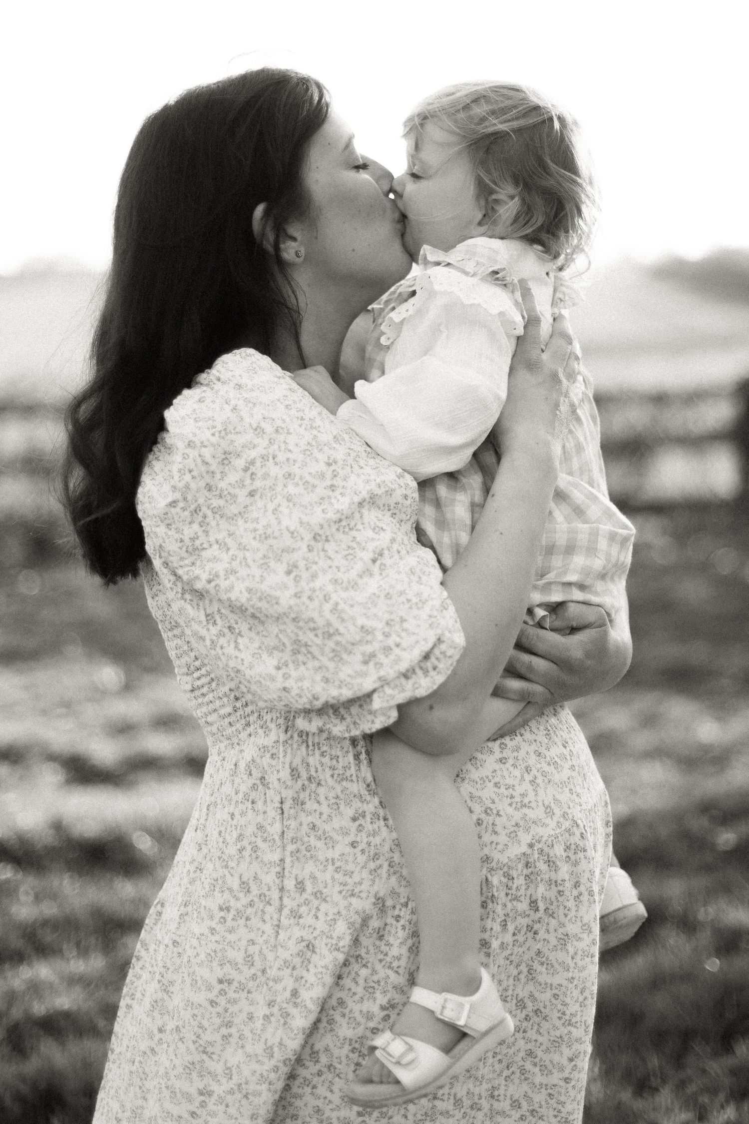Black and white photo of a dark-haired mother sharing a tender kiss with her toddler daughter she is lifting up.