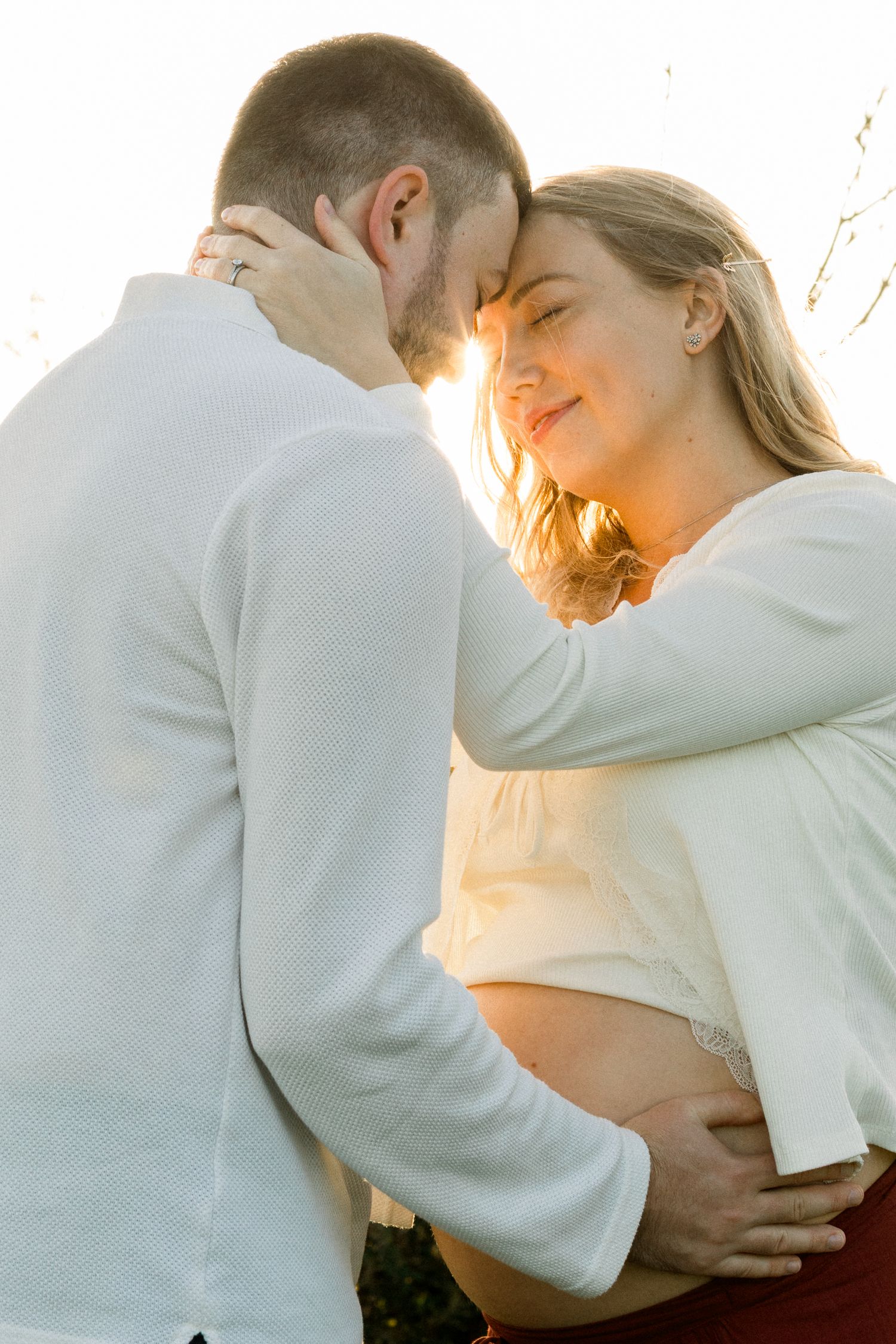 Couple sharing tender moment at golden hour, pregnant woman smiling as man holds her bump outdoors.