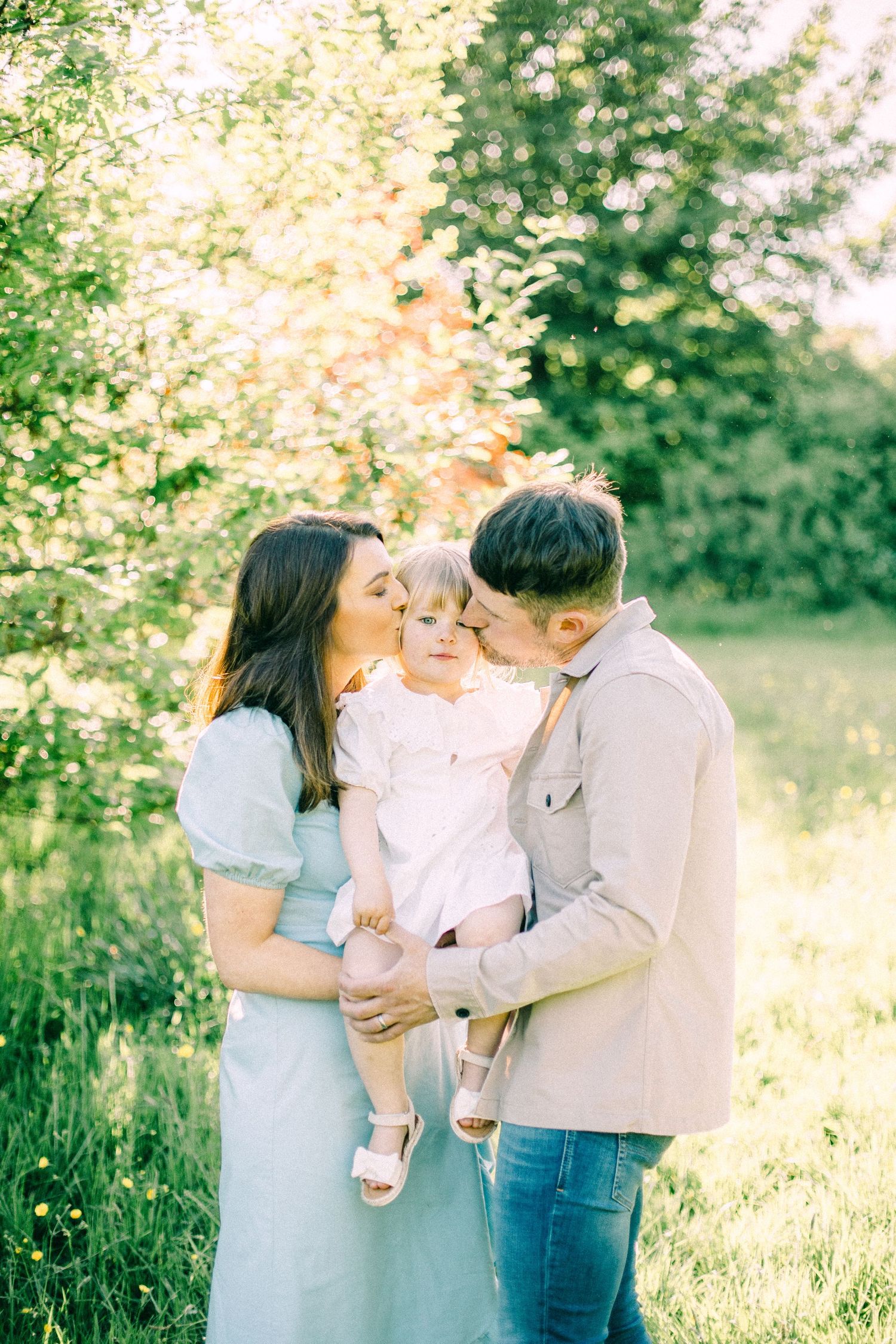 Parents in a sunny green meadow kiss the cheeks of their smiling toddler in a white outfit during a warm family portrait session.