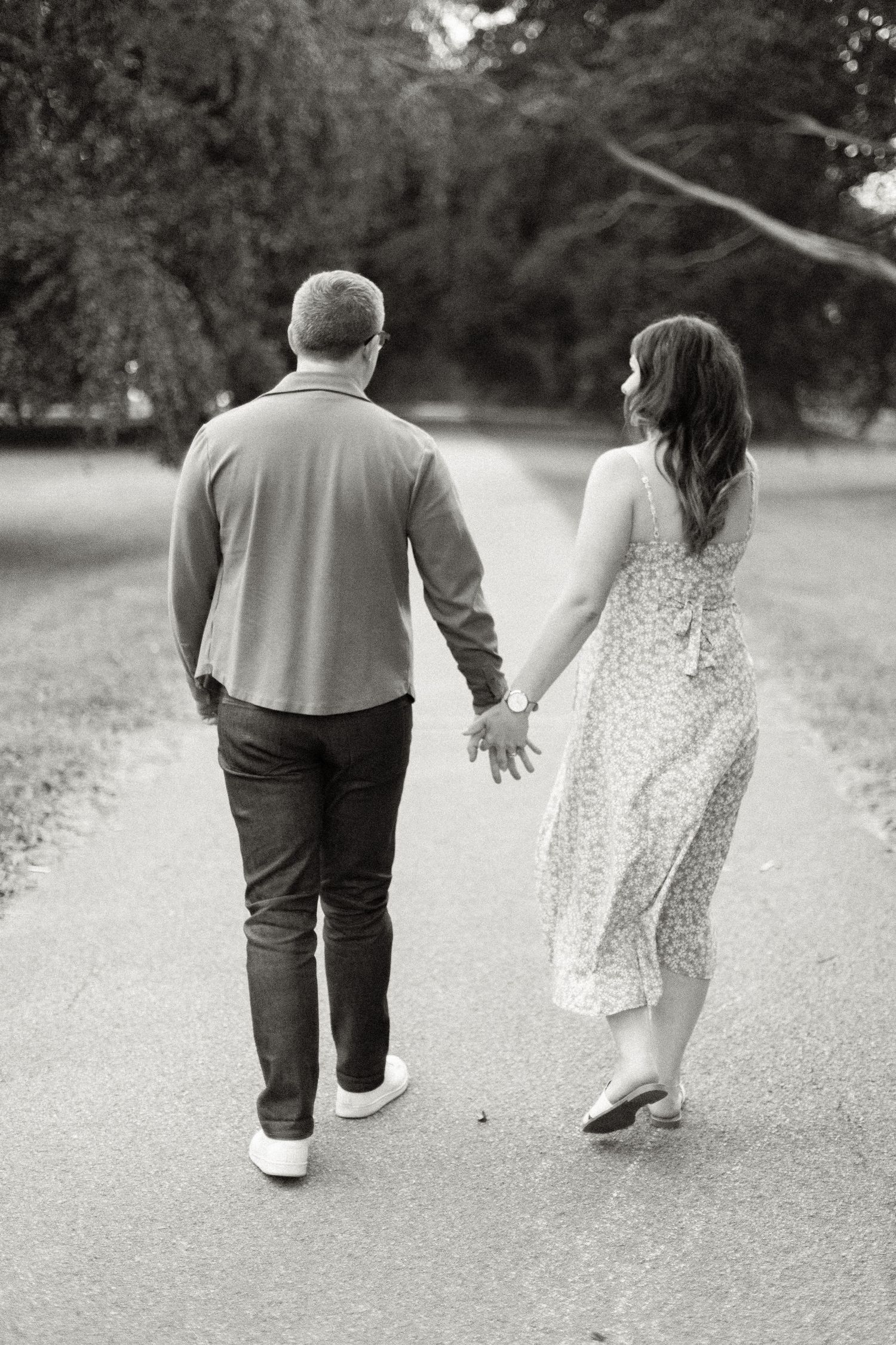 Black and white photo of a couple holding hands walking down a park path, woman in floral dress.
