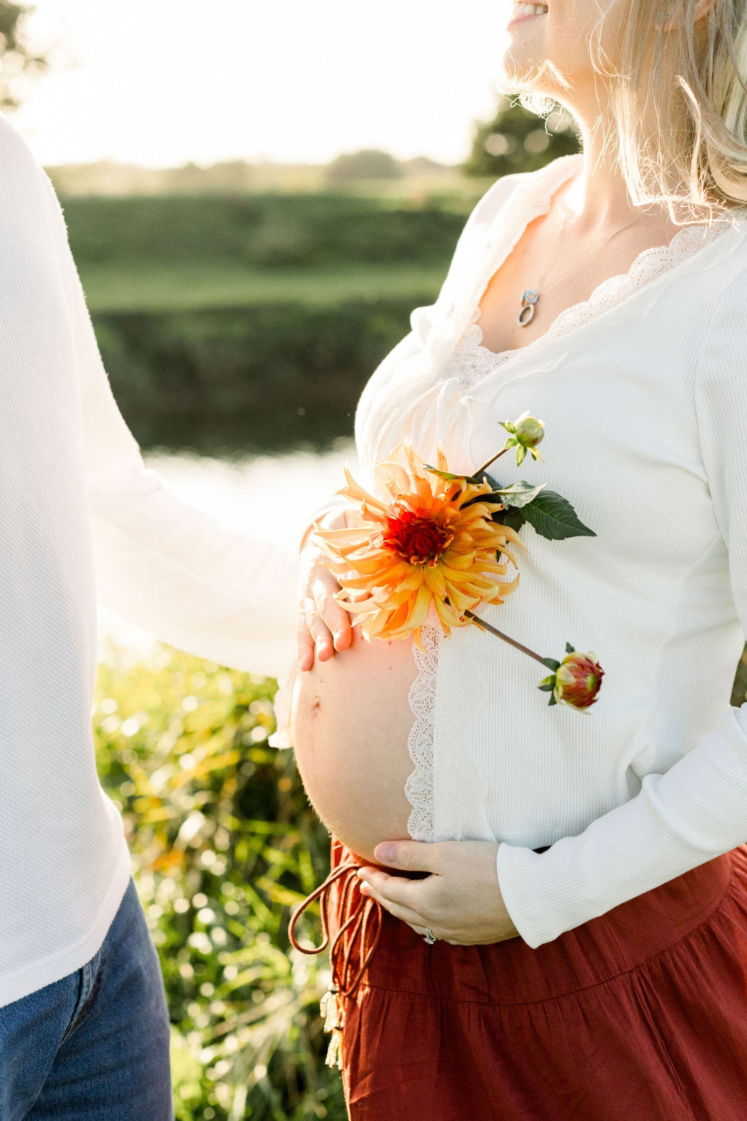 Pregnant woman in white lace top holding orange dahlia flower outdoors near a pond in golden sunlight.