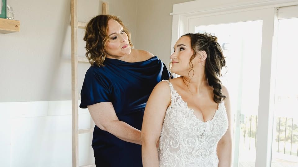 Woman in blue velvet dress adjusting brides lace wedding gown in bright white room.
