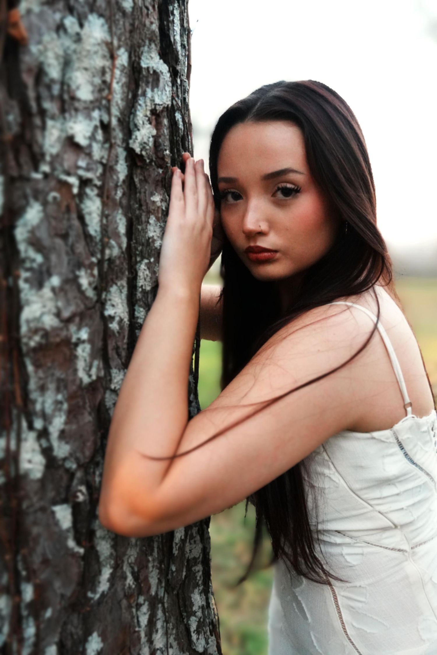 A young brunette woman in a white dress leans gracefully against a birch tree outdoors.