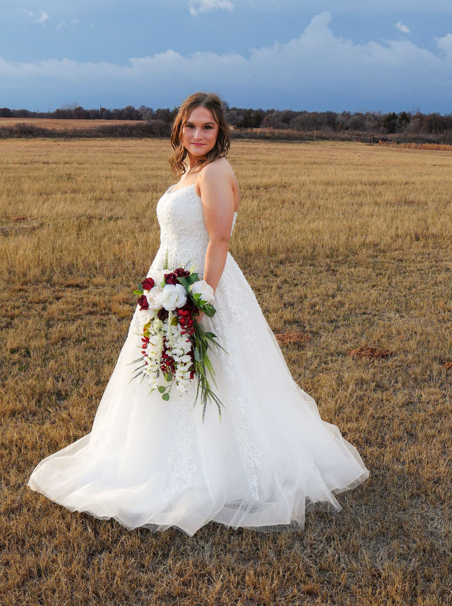 Elegant bride in a white ball gown holding a cascading red and white bouquet in a golden field.