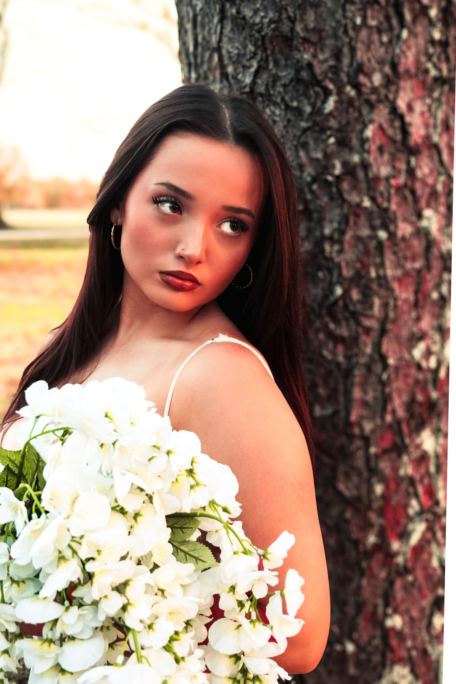 Young brunette woman holding white floral bouquet leaning against tree trunk in warm outdoor lighting.