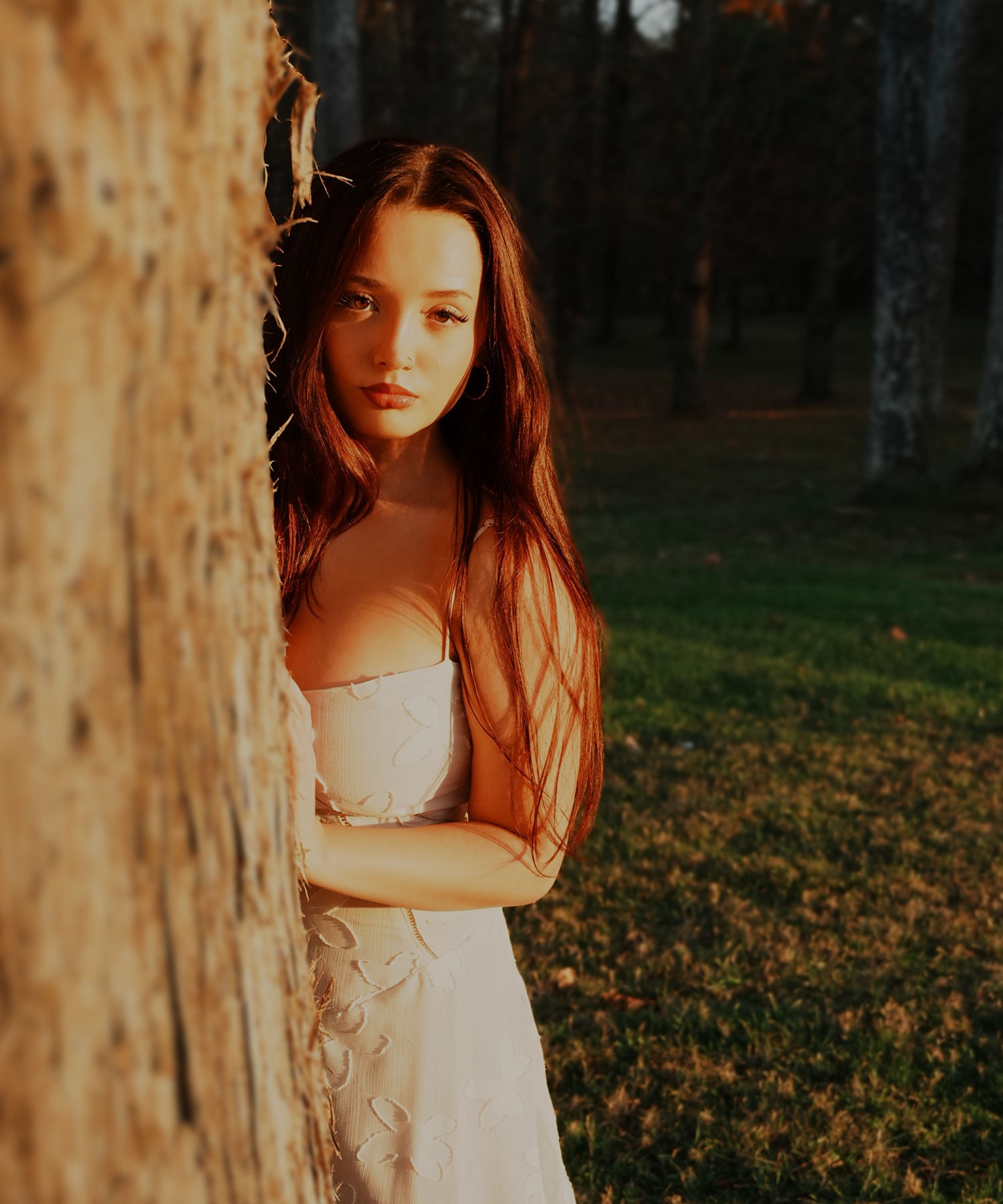 Young woman in white dress leans against a tree in a sun-drenched forest at golden hour.