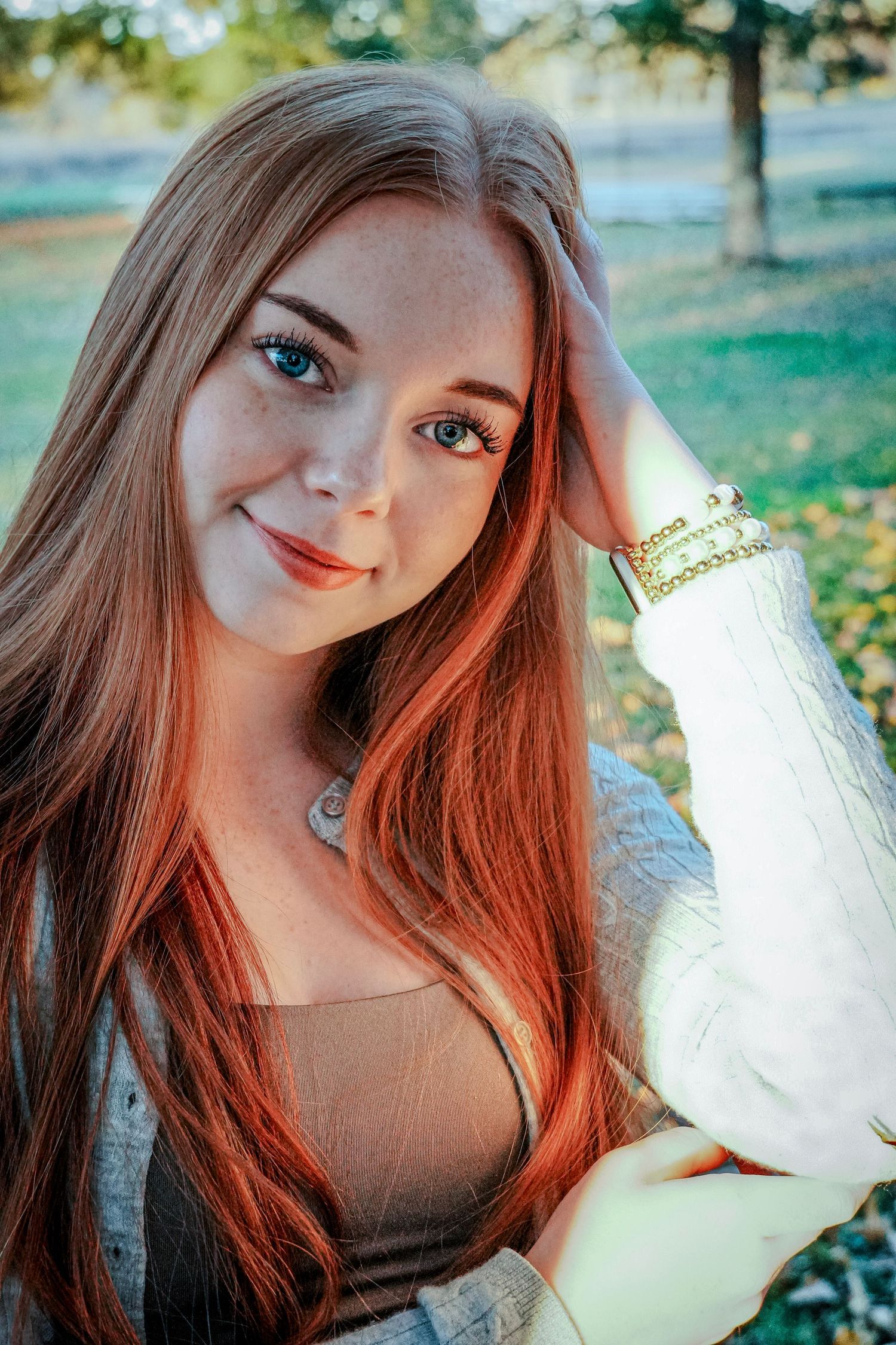 A young woman with long red hair smiles warmly while posing outdoors in natural light.