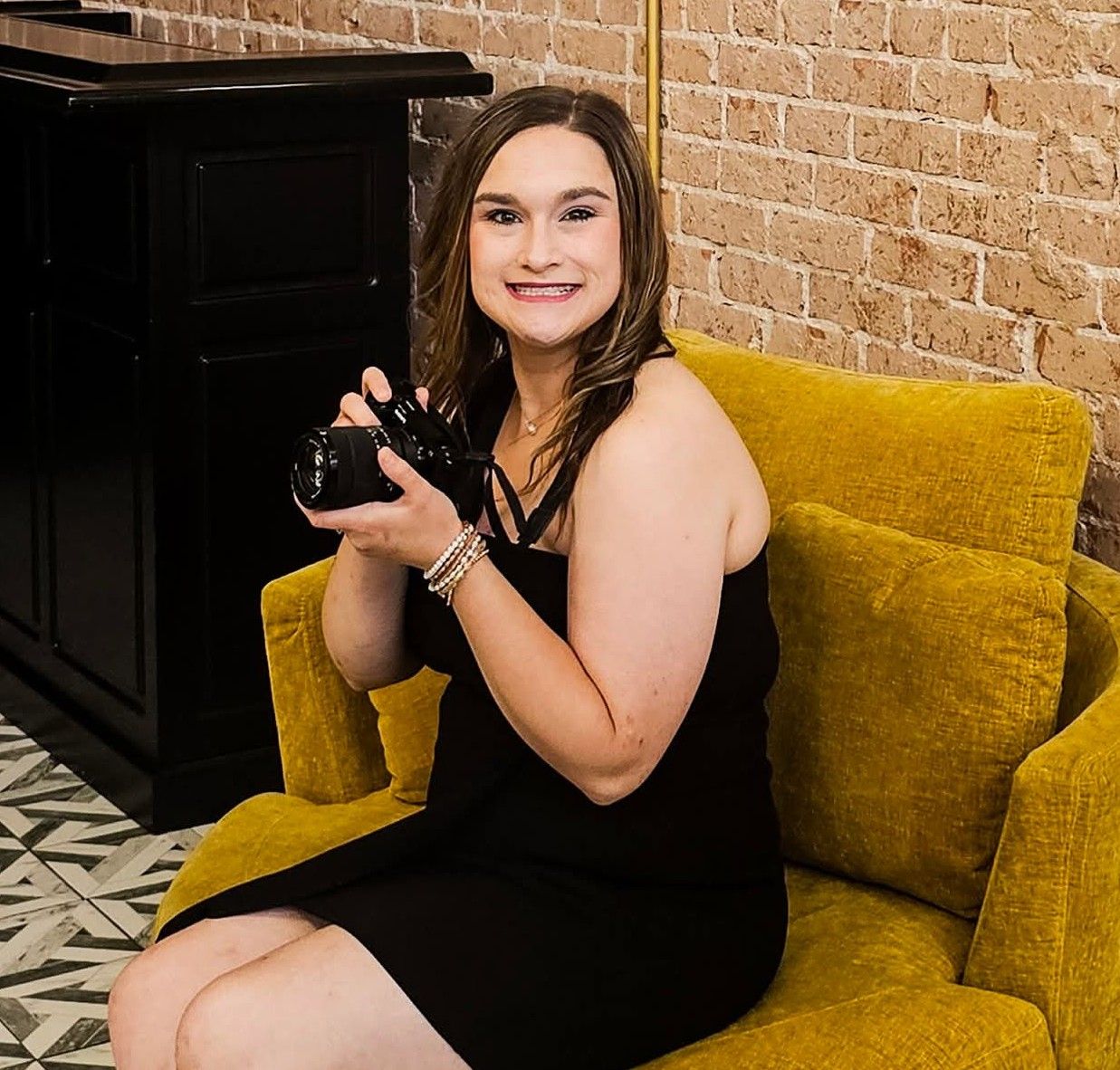 Young woman in black dress holding camera, smiling on yellow velvet chair near brick wall.