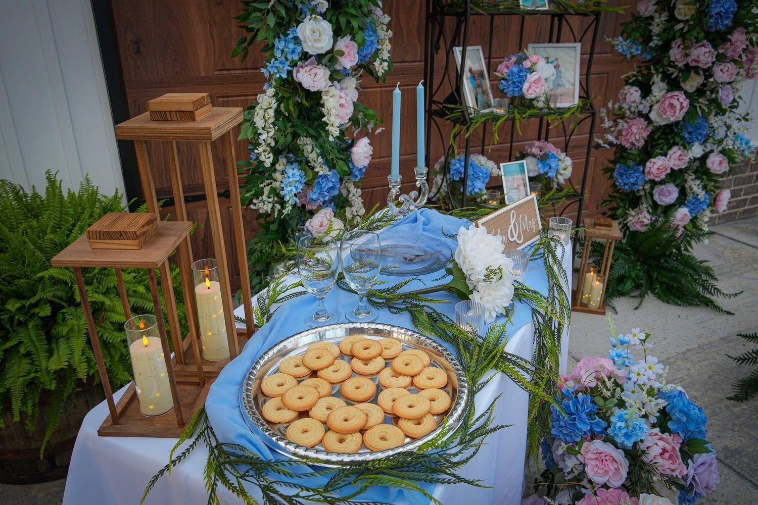 Outdoor wedding welcome table with cookies, candles, lanterns, and pink and blue hydrangea floral decor.