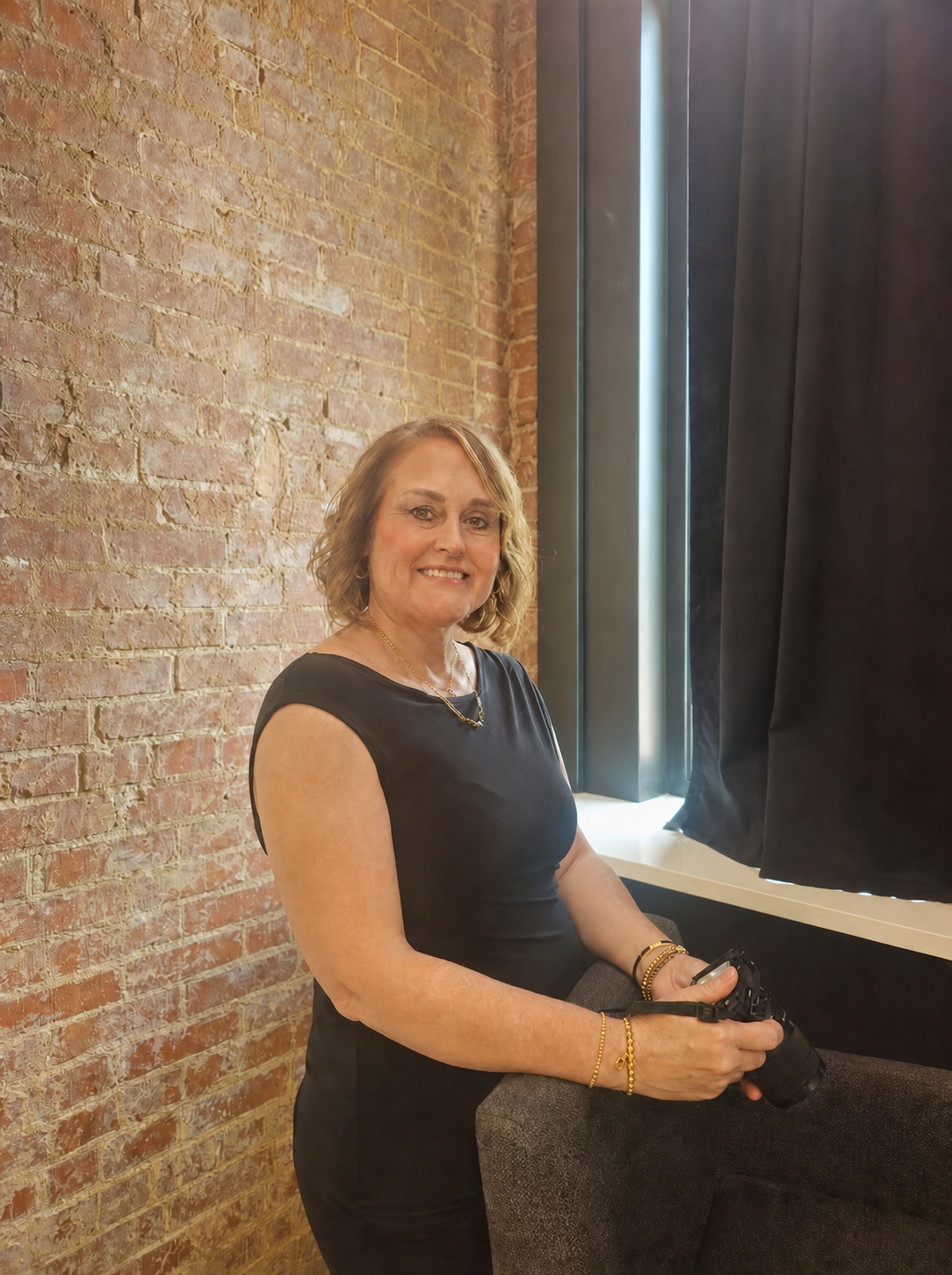 Woman in black dress smiling against exposed brick wall near curtained window holding a camera.
